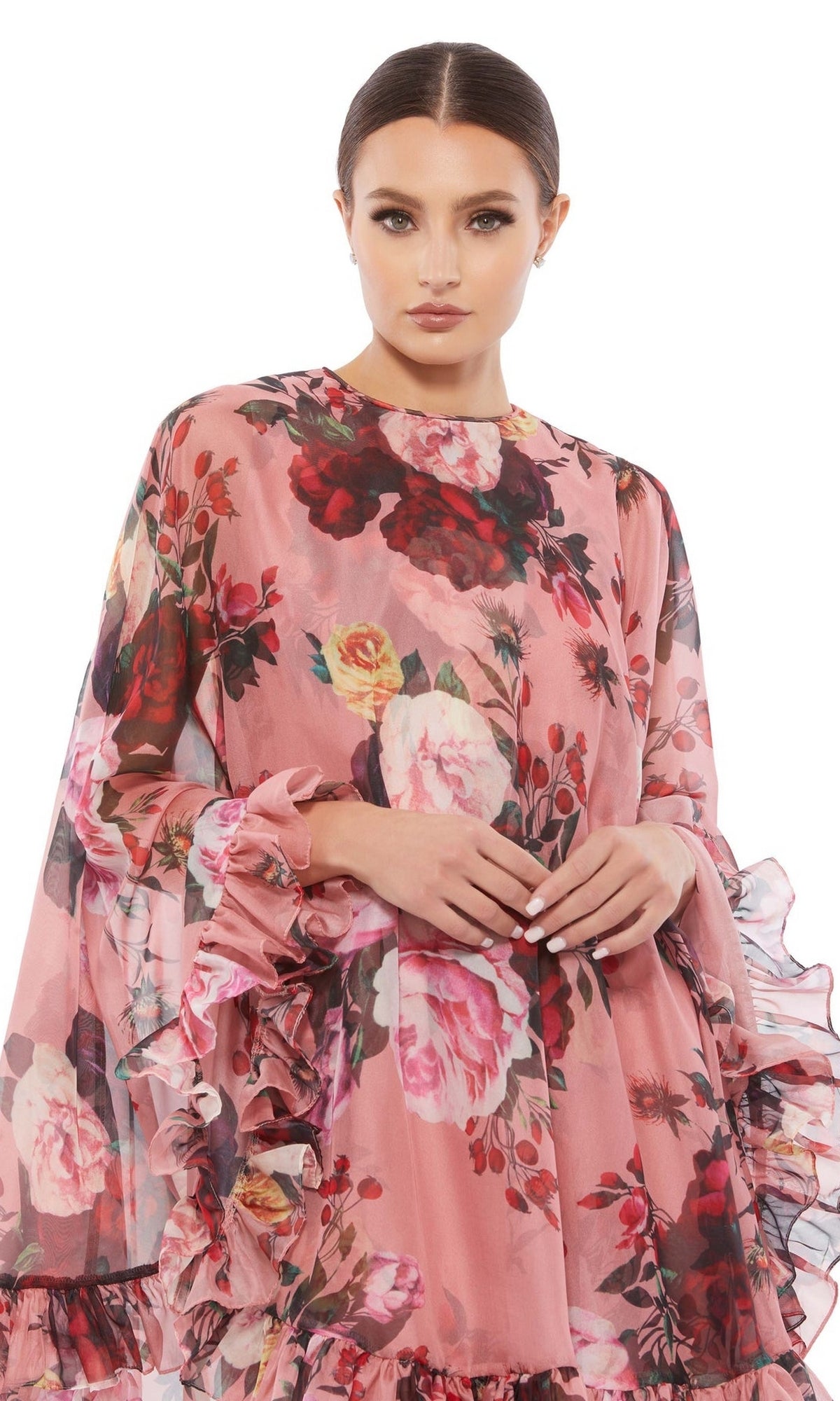 A woman wears the Mac Duggal 55404 Short Print Cape Hoco Dress with pink floral print, ruffles, sheer long sleeves, and cape detail, standing against a plain white background.