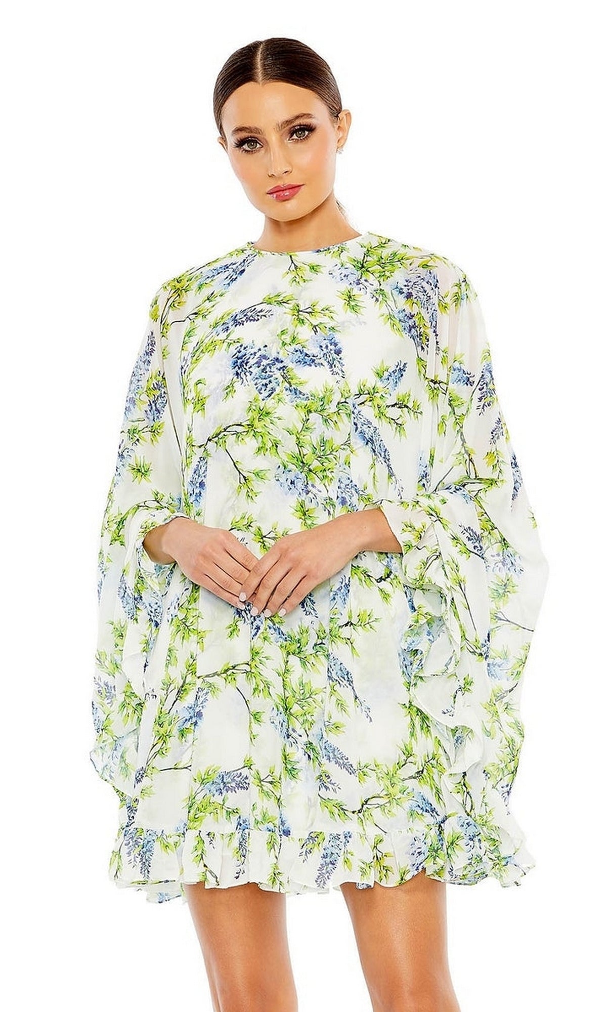 Woman wearing the Mac Duggal 55404 Short Print Cape Hoco Dress— a loose-fitting, long-sleeve white dress with green and purple floral print—stands against a plain white background.