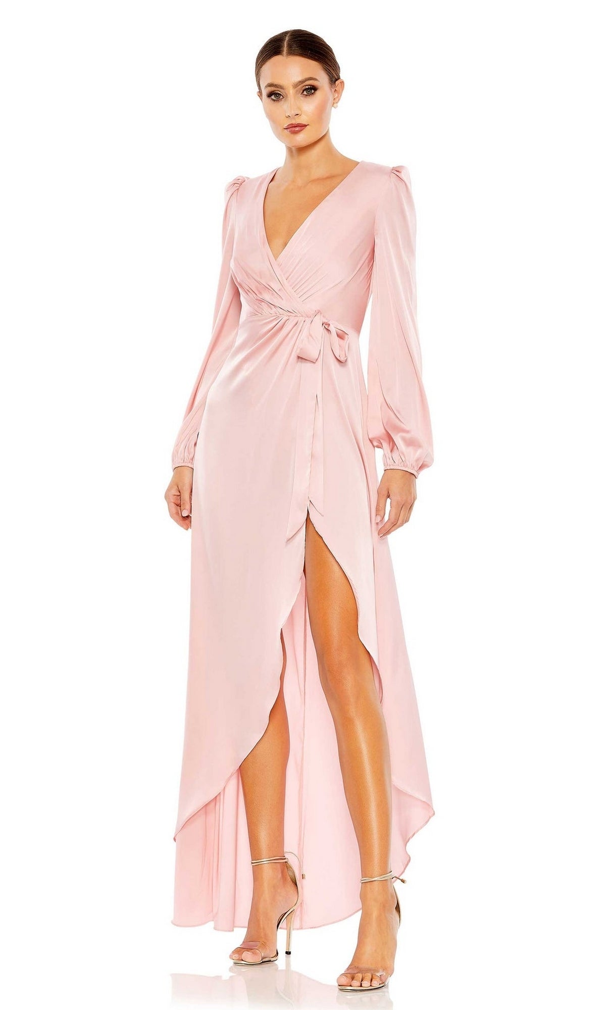 A woman wears the Long Formal Dress 55624 by Mac Duggal in rose pink, styled with a faux wrap design, high slit, v-neckline, and tie waist. She pairs it with silver high-heeled sandals.