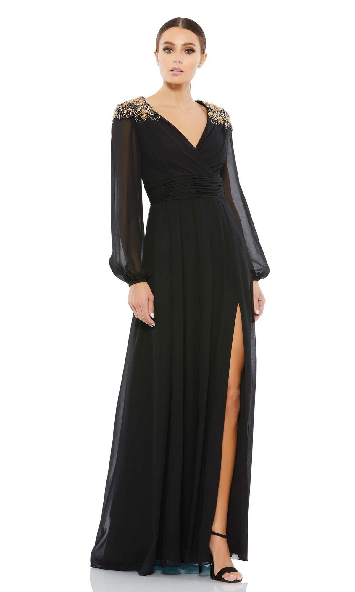 A woman in the Mac Duggal 55694—a black long sleeve chiffon formal dress with sheer sleeves, gold-embellished shoulders, v-neckline, and high leg slit—pairs it perfectly with black high-heeled sandals.