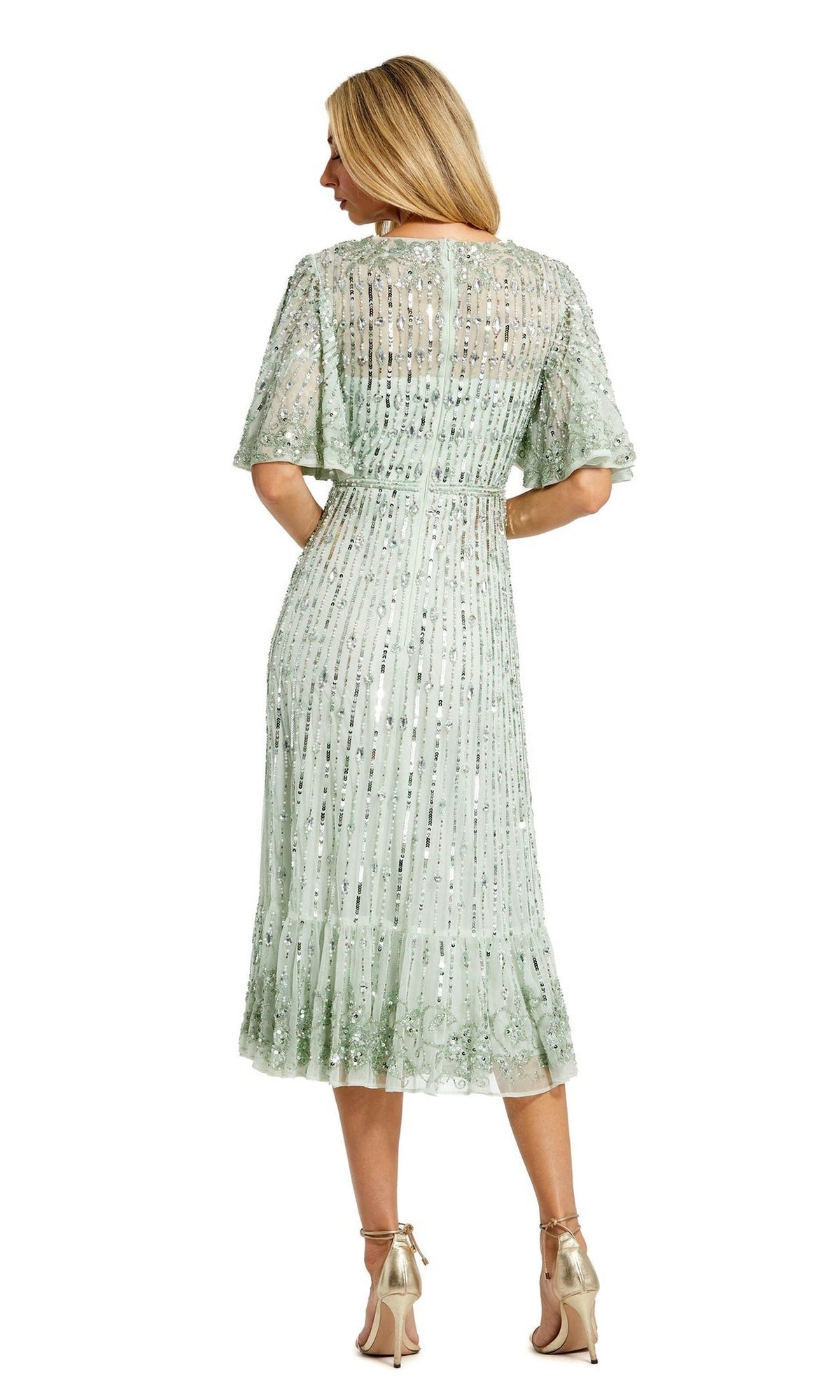 A woman stands facing away, wearing the Mac Duggal 5579 Beaded Midi Semi-Formal Dress in pale green with short sleeves, knee-length hem, and paired with strappy high-heeled sandals.