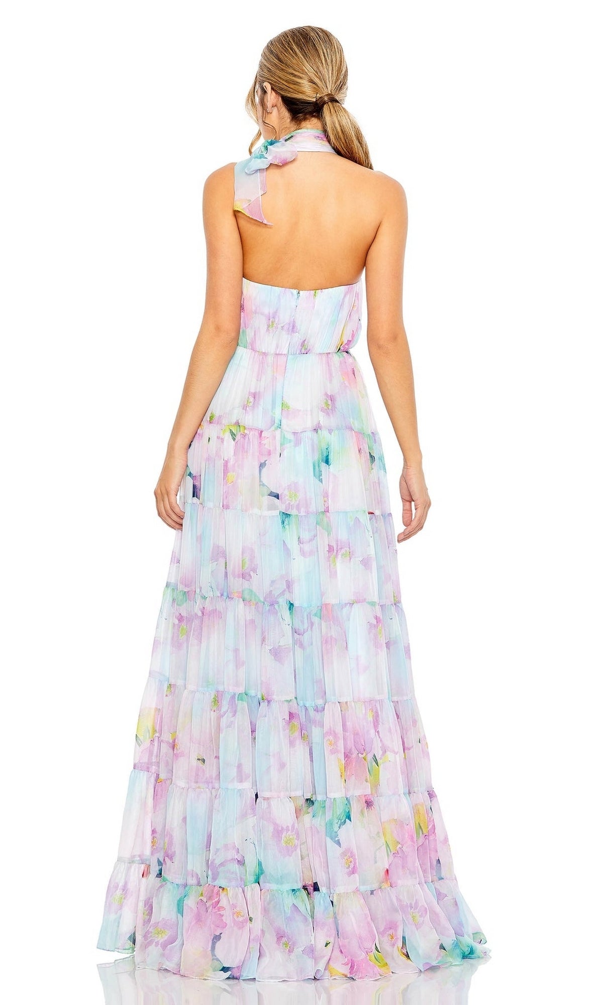 A woman faces away, wearing the Long Formal Dress 55881 by Mac Duggal—a tiered A-line floral print dress with a halter neck and bow detail. Her hair is in a low ponytail.
