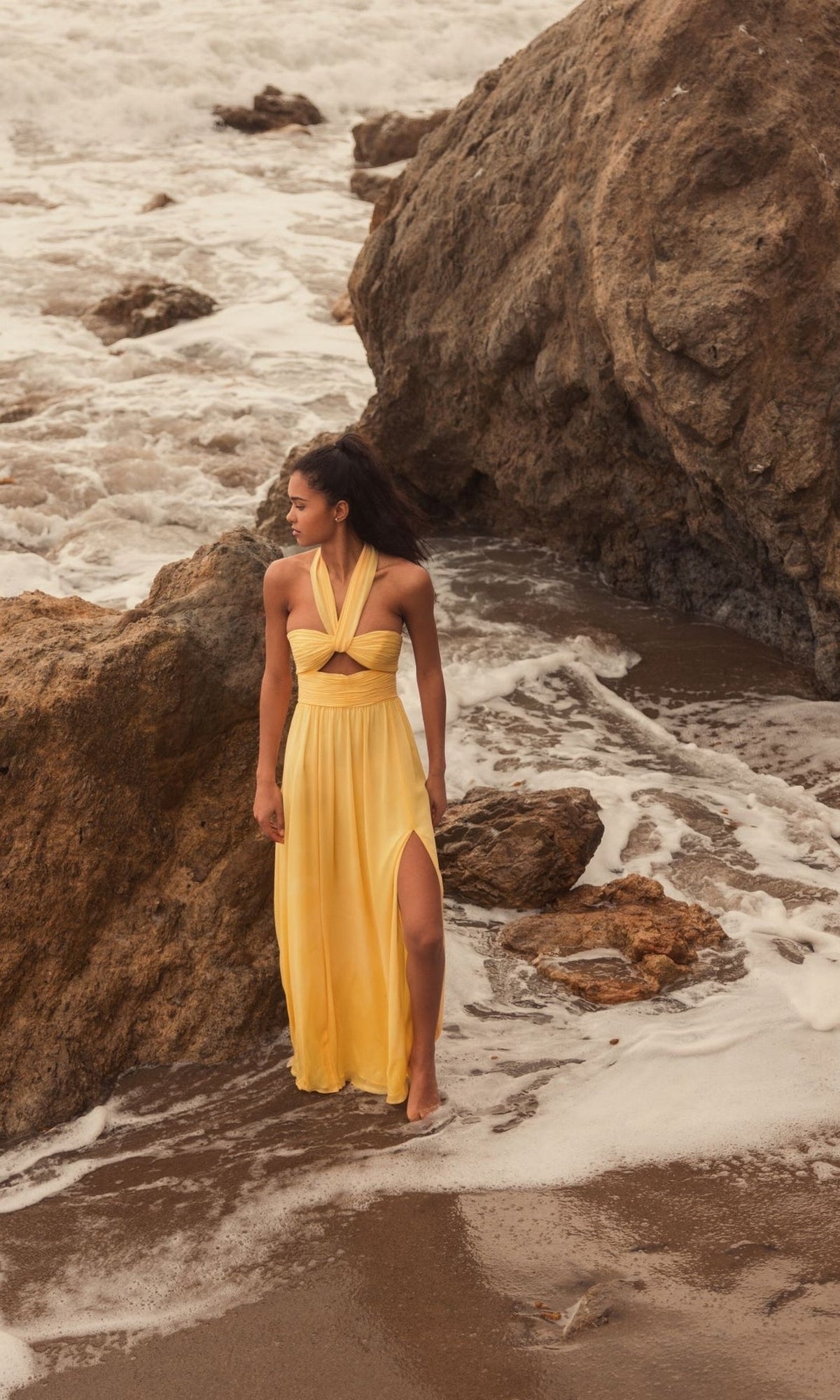 A woman in the Long Formal Dress 55898 by Mac Duggal stands barefoot on wet sand near large rocks as waves wash ashore.