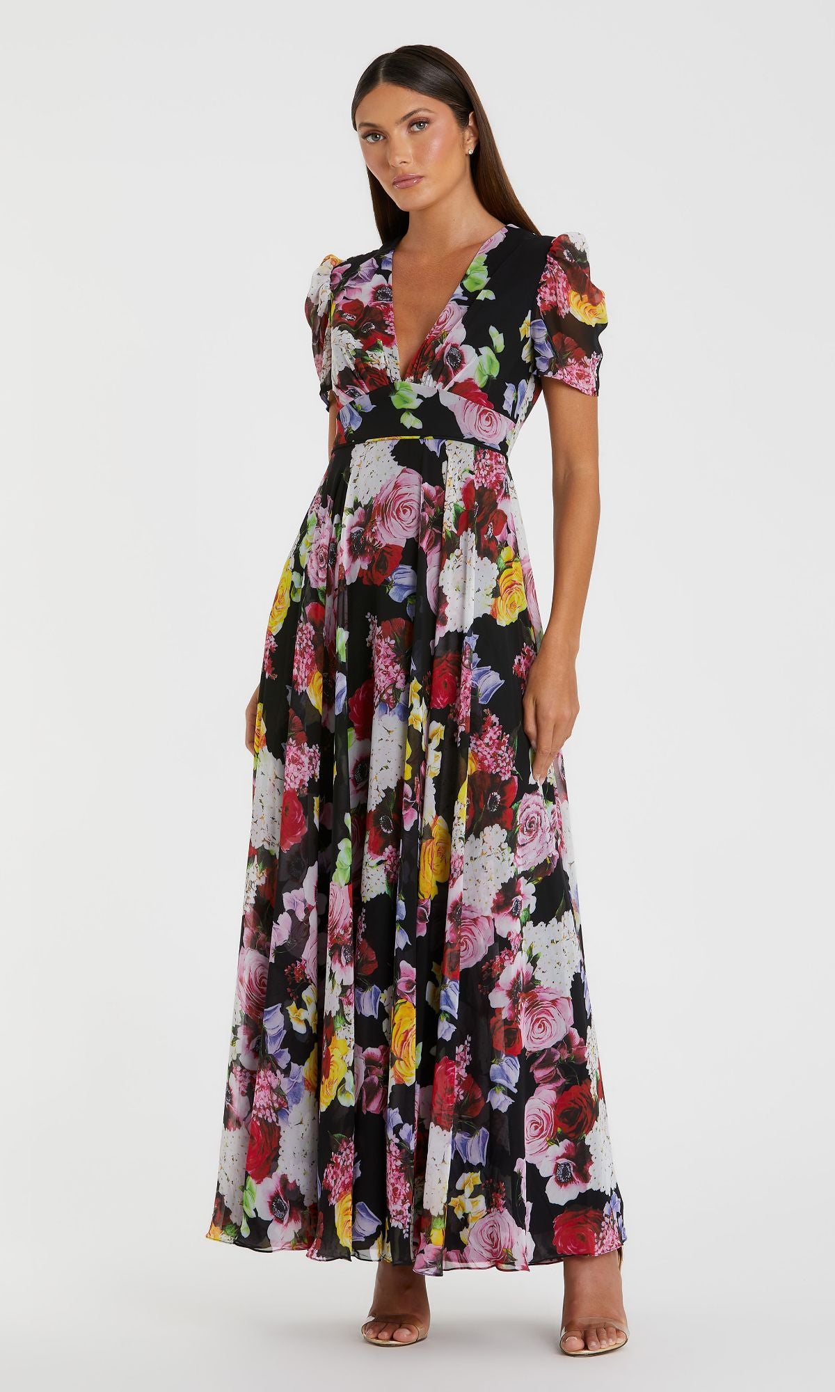 A woman wears the Mac Duggal 55933 Short Sleeve Print Formal Dress, featuring a deep V-neck and colorful floral print, paired with open-toe heels.