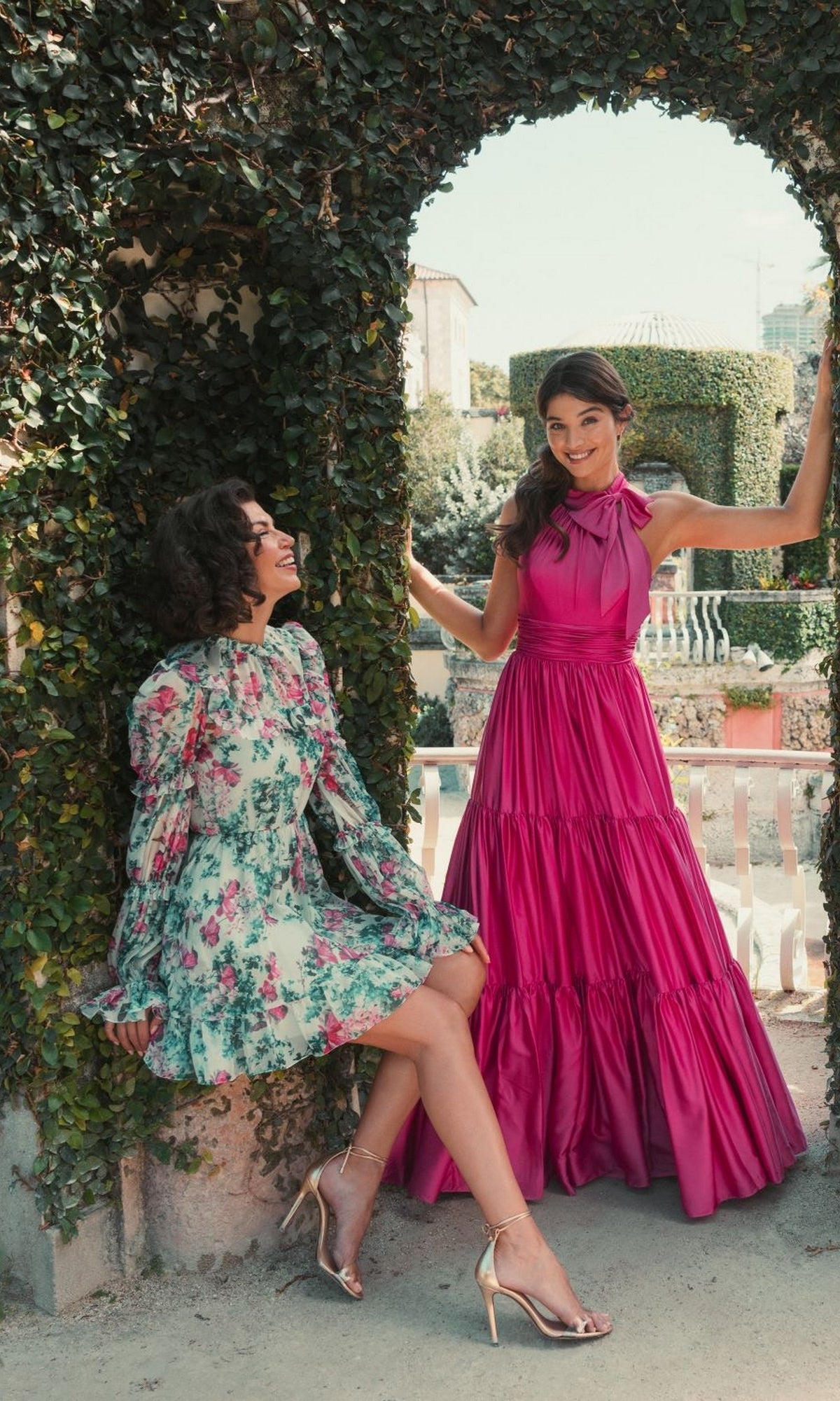 Two women pose beneath an ivy-covered stone arch; one smiles in the Mac Duggal High-Neck Print Party Dress 56008, while her friend stands beside her in heels and a long-sleeve dress as they enjoy the garden setting.