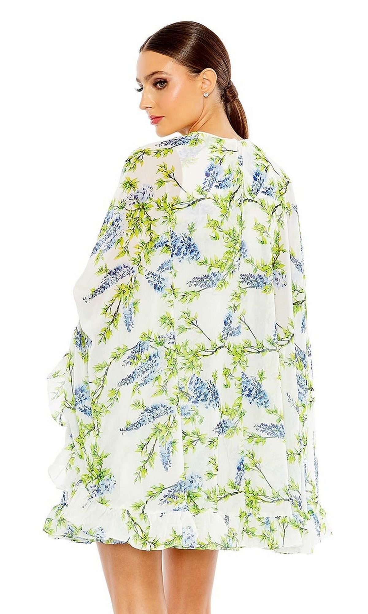 A woman with brown hair in a low ponytail is seen from the back wearing the Mac Duggal Cape Short Party Dress 56029, a loose white dress with blue and green floral designs.