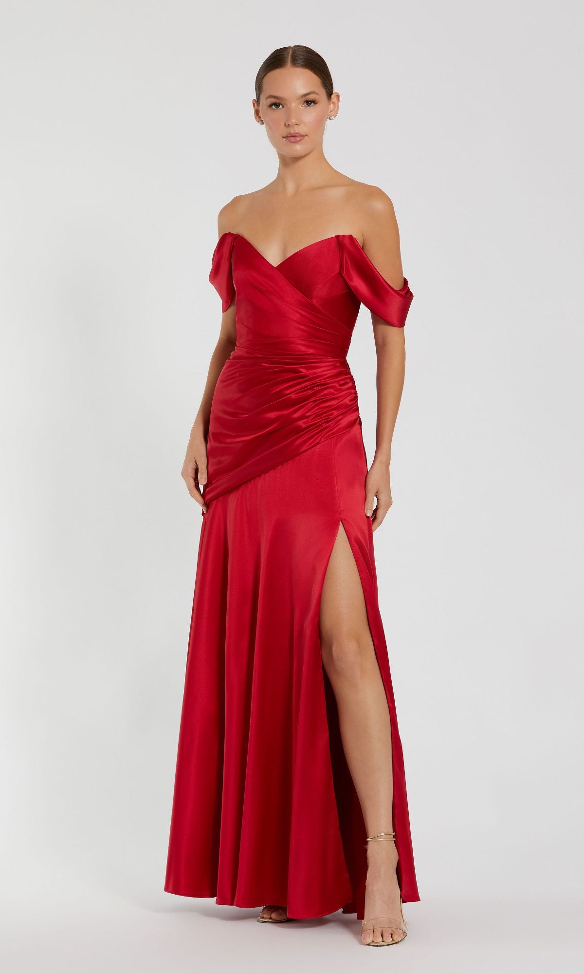 A woman wears the Mac Duggal 56037 off-the-shoulder prom dress in red satin, featuring a thigh-high slit, as she stands against a plain light background.
