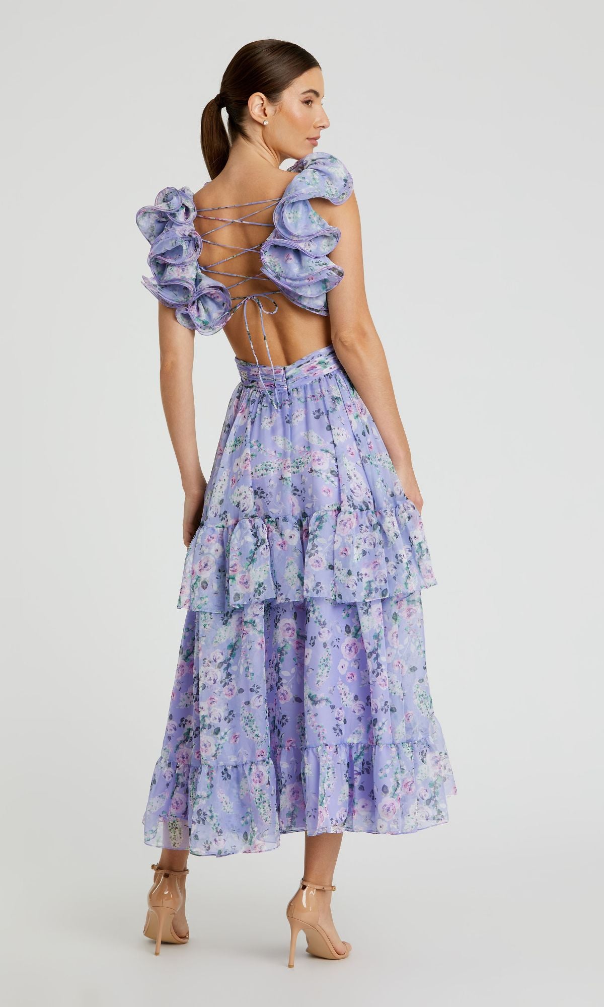 A woman stands in beige heels, facing away, wearing the Mac Duggal 56049 Midi Lilac Print Party Dress featuring floral print, ruffled details, an open back, and lace-up accents.