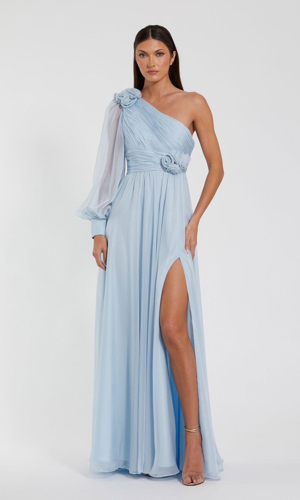 A woman wearing the Mac Duggal 56070 light blue long formal dress with a one-sleeve design, sheer long sleeve, floral accents, and high slit stands against a plain white background.