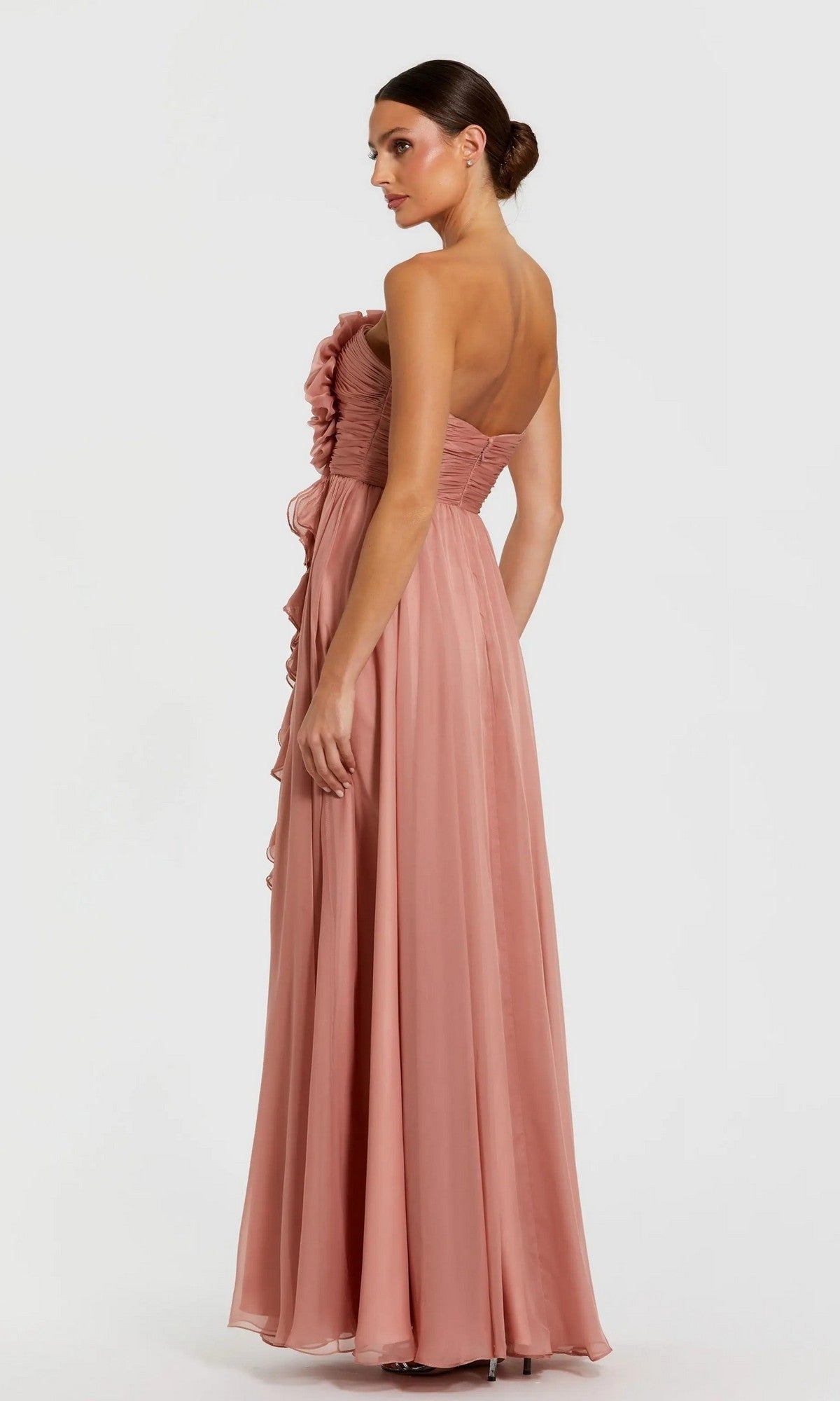 A woman in the Mac Duggal 56071 long formal dress—featuring a strapless, floor-length silhouette in dusty rose, with ruffle details and a delicate 3D floral bodice—stands against a plain background, looking over her shoulder.