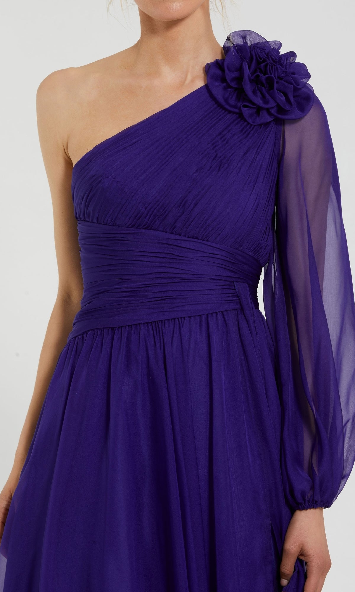 A woman wears the Mac Duggal 56073 One-Shoulder Purple Formal Dress, featuring an indigo purple hue, sheer long sleeve, and a large fabric flower on the shoulder.