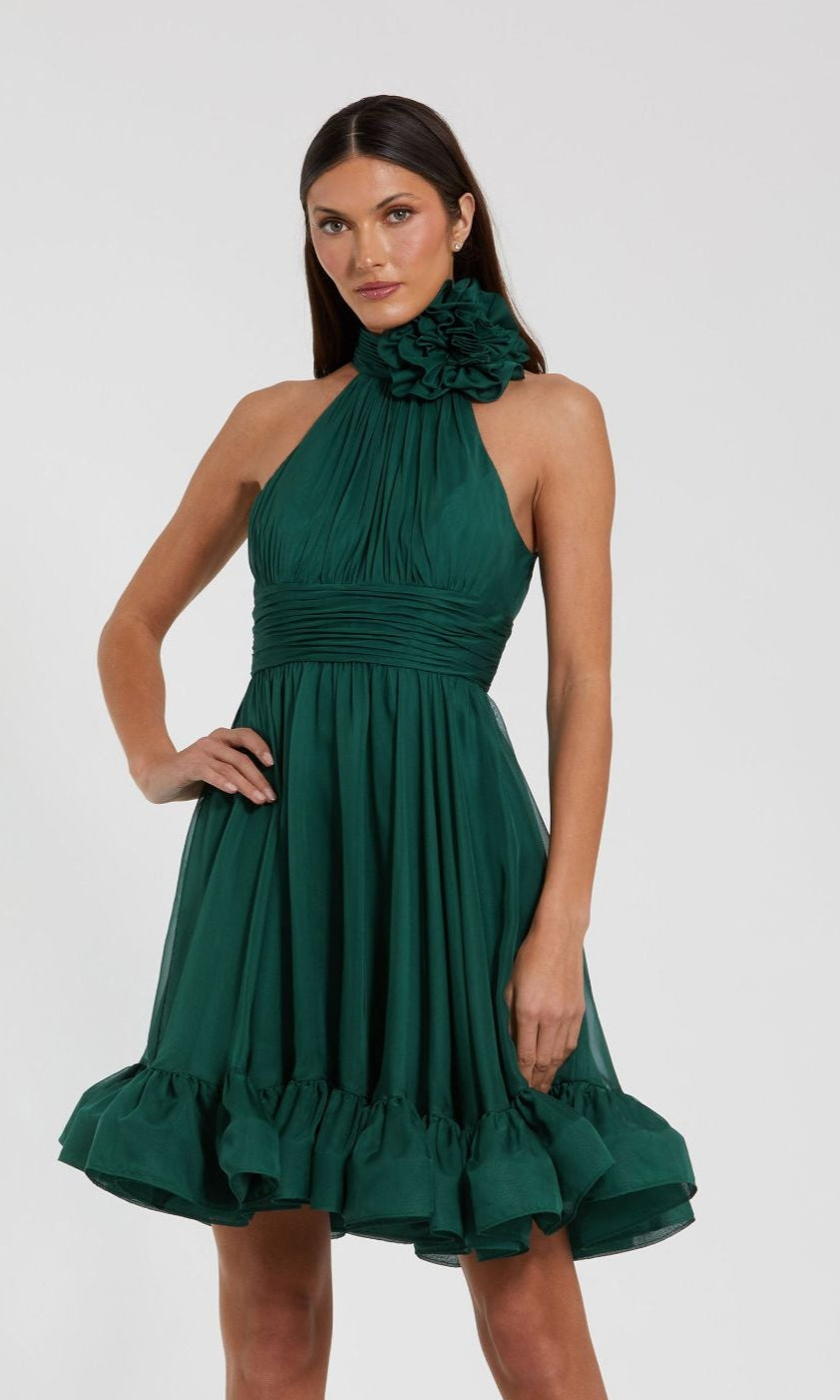 A woman wears the Short Wedding Guest Dress 56080 by Mac Duggal, a sleeveless teal green dress with a halter neckline, large ruffle accent, and ruffled hem, standing against a plain light background.