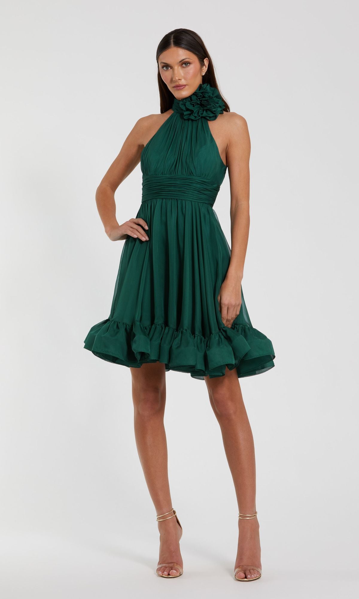 A woman wears the Short Wedding Guest Dress 56080 by Mac Duggal, featuring a sleeveless teal green halter neckline, floral neck detail, and ruffled hem, standing against a plain light background.