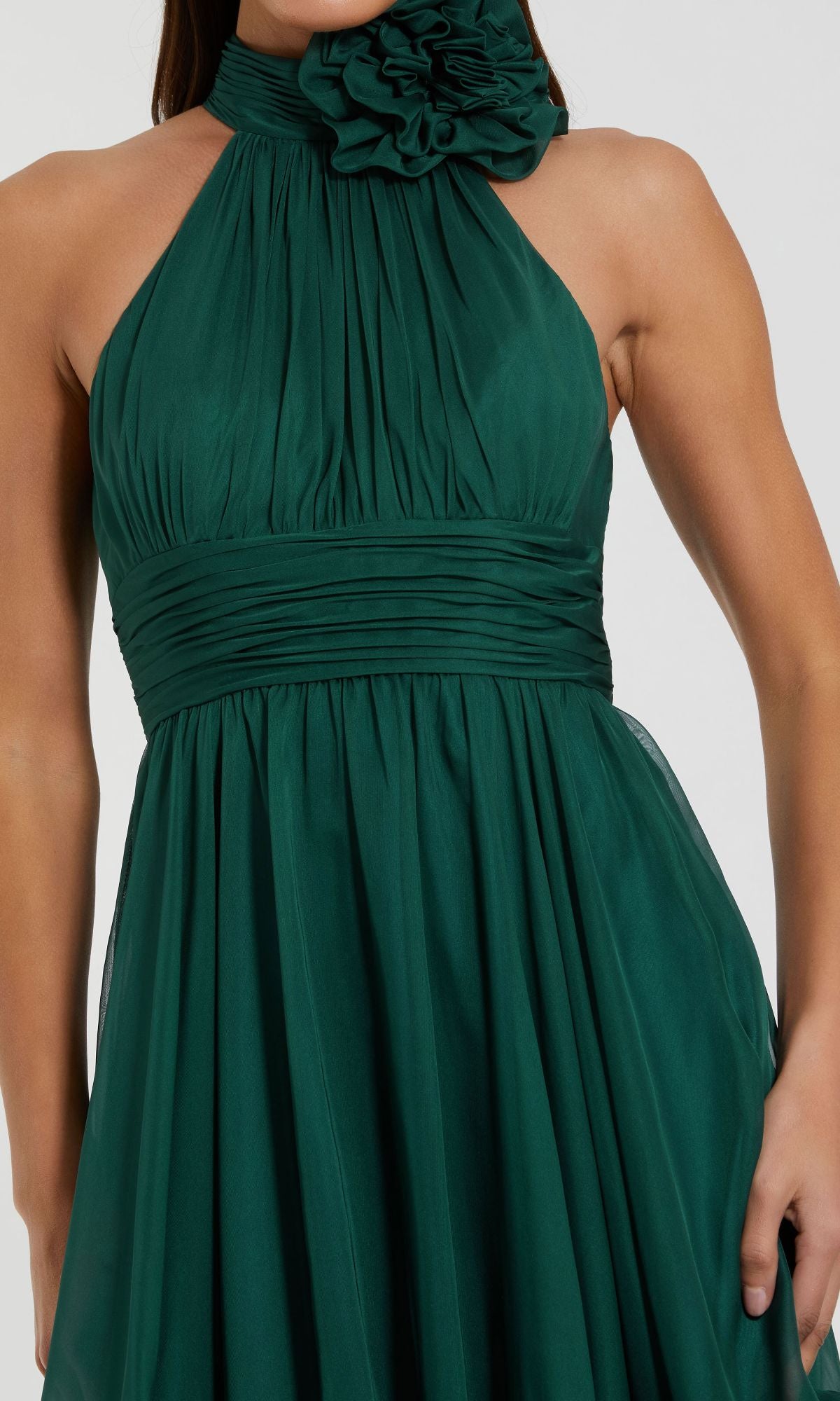 A woman in the Mac Duggal Short Wedding Guest Dress 56080, featuring a sleeveless teal green design with pleated bodice, wide waistband, halter neckline, and large fabric flower detail at the neck.