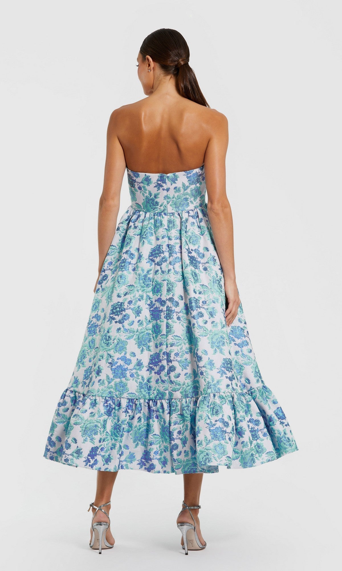 Model facing away in the Mac Duggal 56092 Midi Wedding Guest Dress, featuring a strapless blue and green floral pattern with a ruffled hem, styled with silver heels—an elegant pick for weddings.