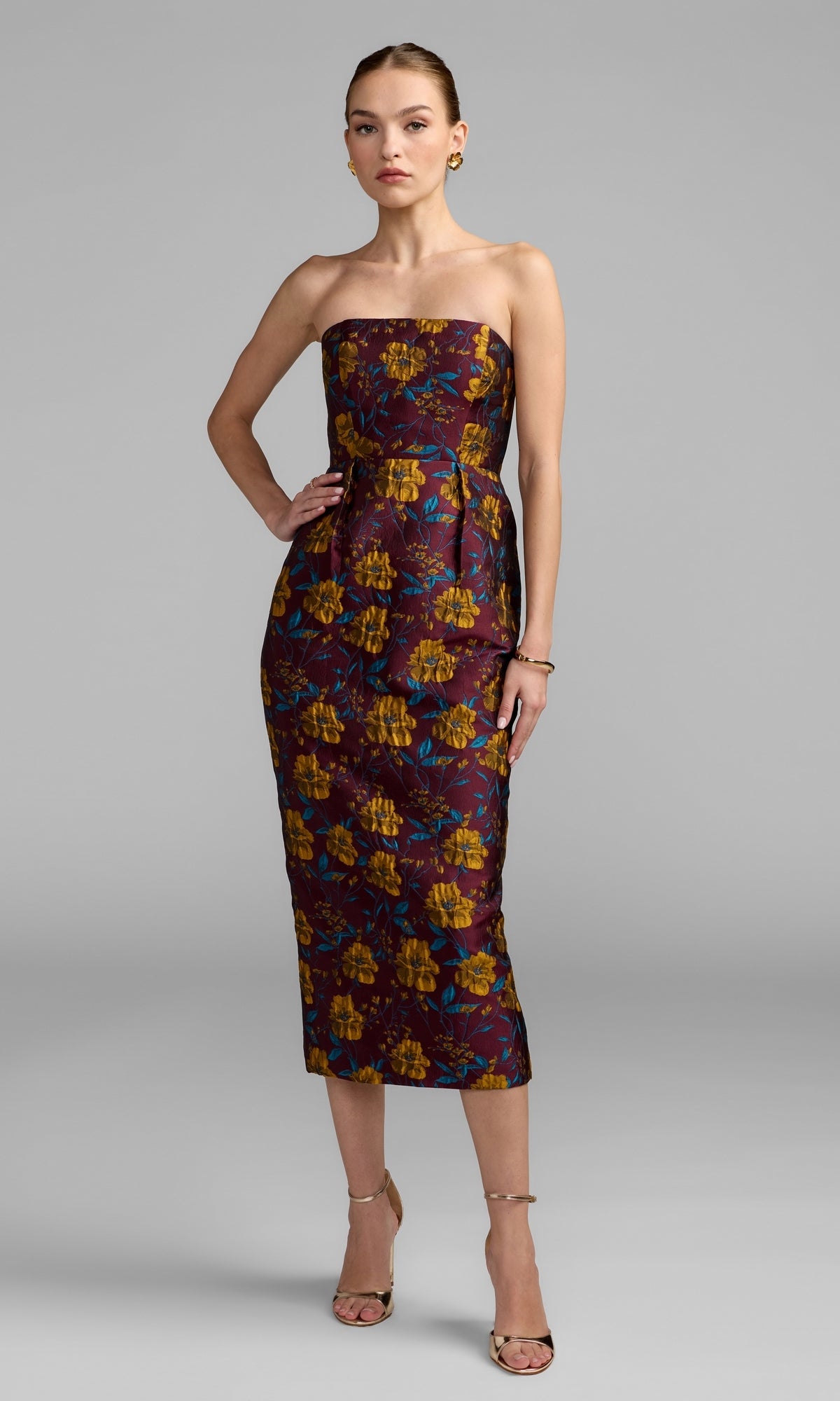 A woman in the Mac Duggal 56097 Midi Floral Wedding Guest Dress in Plum Multi stands against a plain gray background, paired with gold sandals and minimal jewelry—an elegant choice for a wedding guest ensemble.