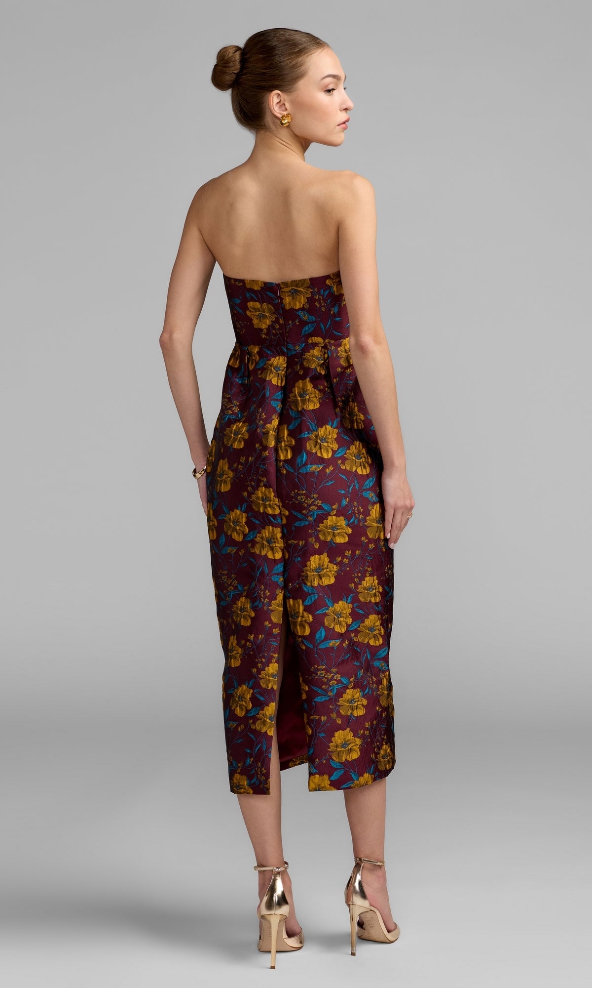 Woman in a strapless, maroon floral-print Midi Floral Wedding Guest Dress: Mac Duggal 56097 with gold heels, standing on a gray background, facing away from the camera.