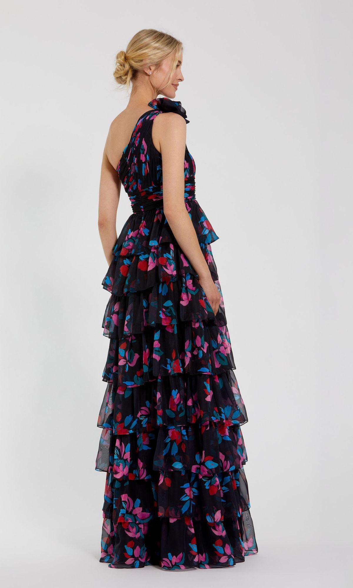 A woman in the Mac Duggal 56120 Long Print Ruffled Formal Dress—black, sleeveless, tiered, with colorful floral patterns—stands sideways against a plain light background.