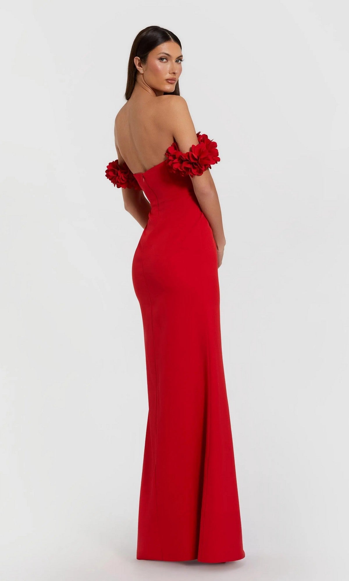 A woman stands against a light background, looking over her shoulder in the Mac Duggal 56131 Floral-Straps Long Formal Dress—a fitted, floor-length red gown with floral off-the-shoulder straps.