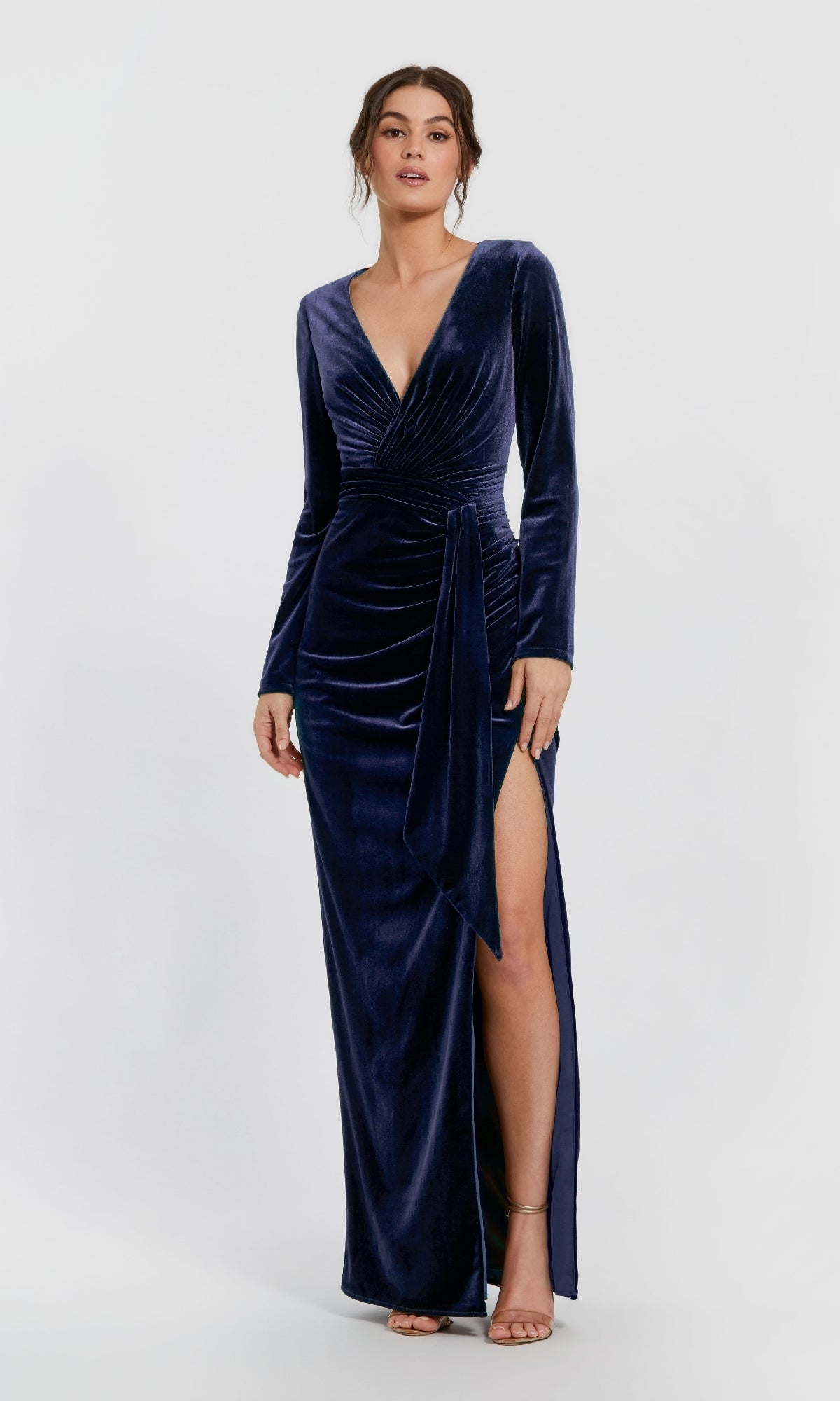 A woman models the Mac Duggal 56143 long formal dress, featuring deep blue velvet, long sleeves, a deep V-neckline, ruched waist, and high slit against a light background.