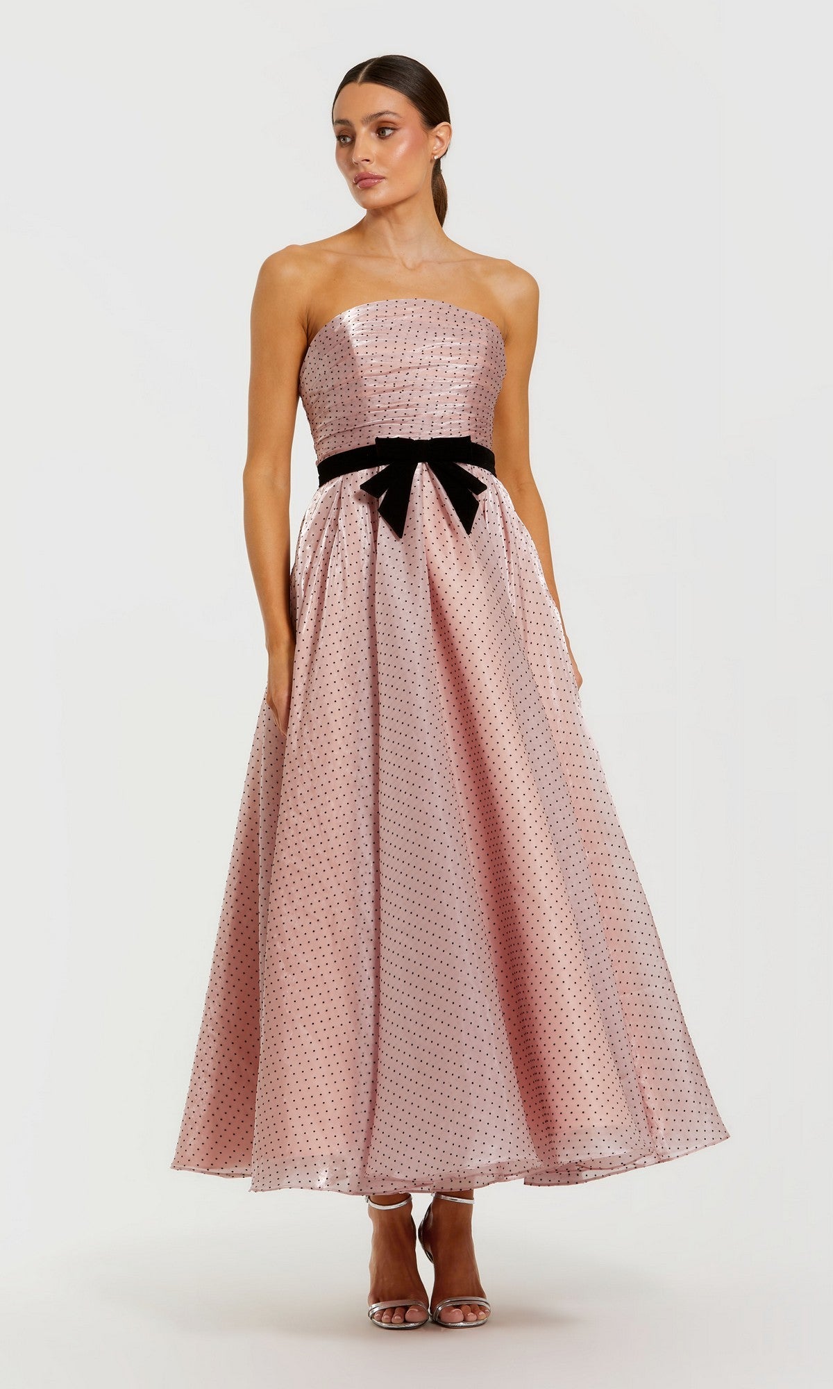 A woman wears the Mac Duggal 56184 long formal dress—a strapless, rose pink organza gown with polka dots and a black bow at the waist—standing against a plain light background.