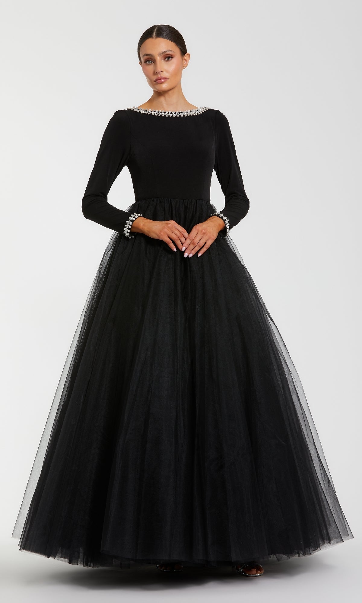 A woman poses against a white background, wearing the Mac Duggal 56186โa long sleeve black formal gown with pearl-detailed neckline and cuffs, and a full tulle skirt for an elegant evening look.