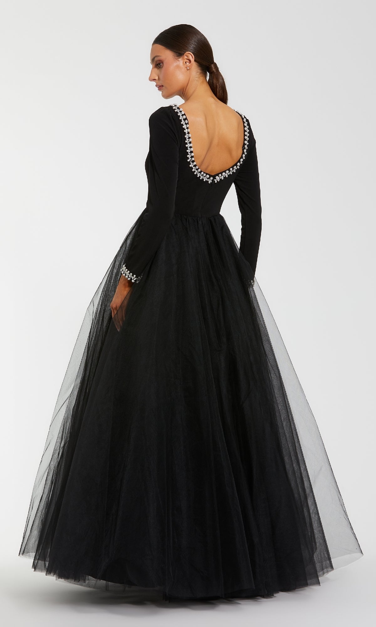 A woman poses in the Mac Duggal 56186, a long sleeve black formal gown featuring a tulle skirt, deep scoop back, and pearl accents on the neckline and cuffs.