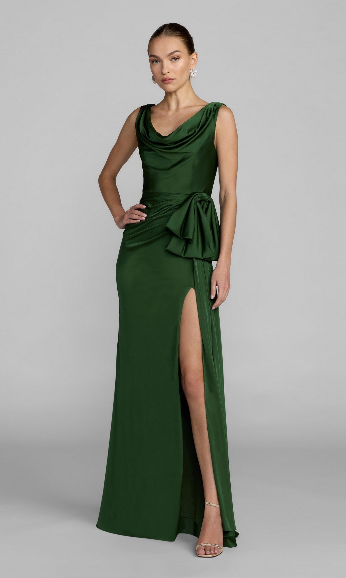 A woman wearing the Mac Duggal 56230 Long Formal Dress in emerald—a sleeveless, floor-length charmeuse gown featuring a cowl neckline, side bow detail, and a high slit—stands against a plain gray background.