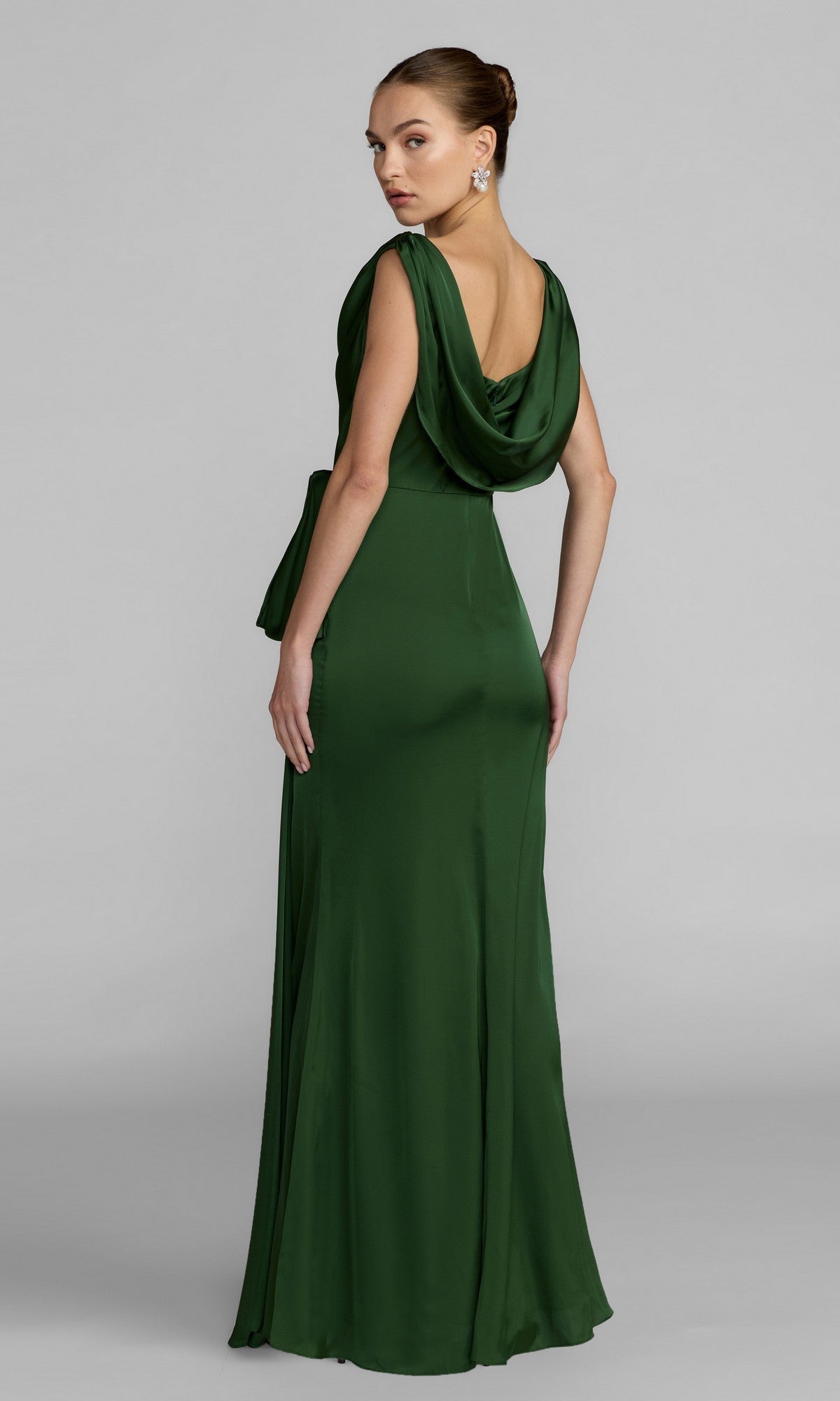 A woman stands on a plain gray background, looking over her shoulder while wearing the Mac Duggal 56230—a floor-length, sleeveless green charmeuse formal dress featuring a draped back and a subtle cowl neckline.