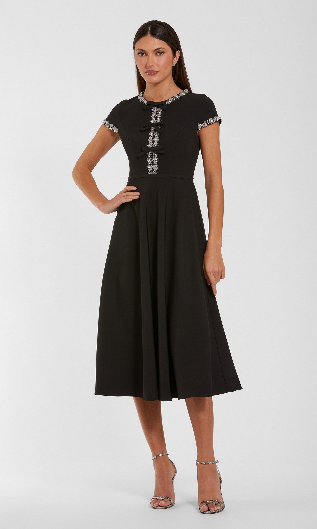 A woman poses against a plain background in the Mac Duggal 56242 Midi Party Dress, a black, short-sleeved, knee-length design featuring decorative detailing on the sleeves and front. Paired with silver high-heeled sandals, it’s an elegant option for wedding guests or as a sophisticated midi party dress.