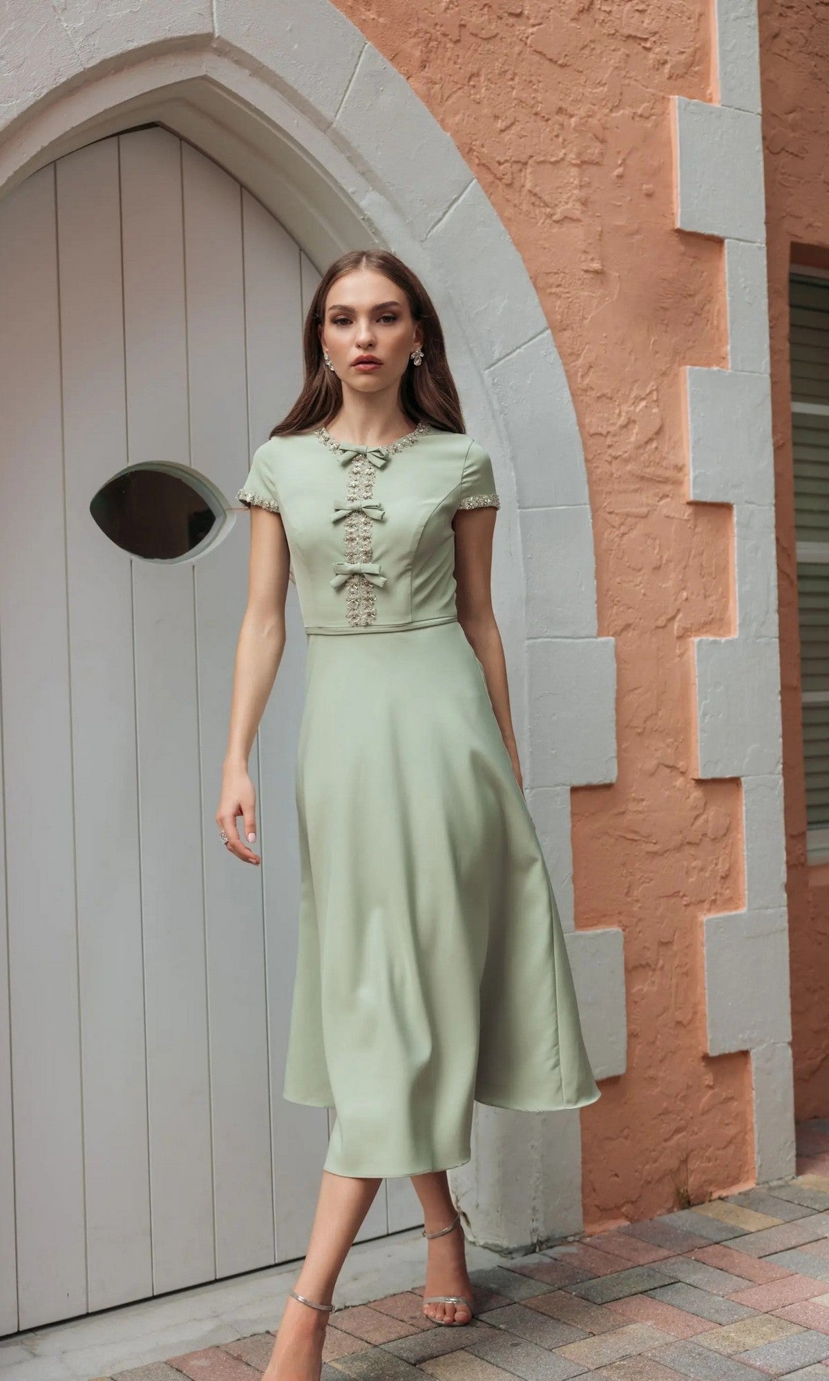 A woman in the Mac Duggal 56242 Midi Party Dress in sage, featuring short sleeves and lace detailing, walks on a brick path near an arched white door and a textured peach wall.