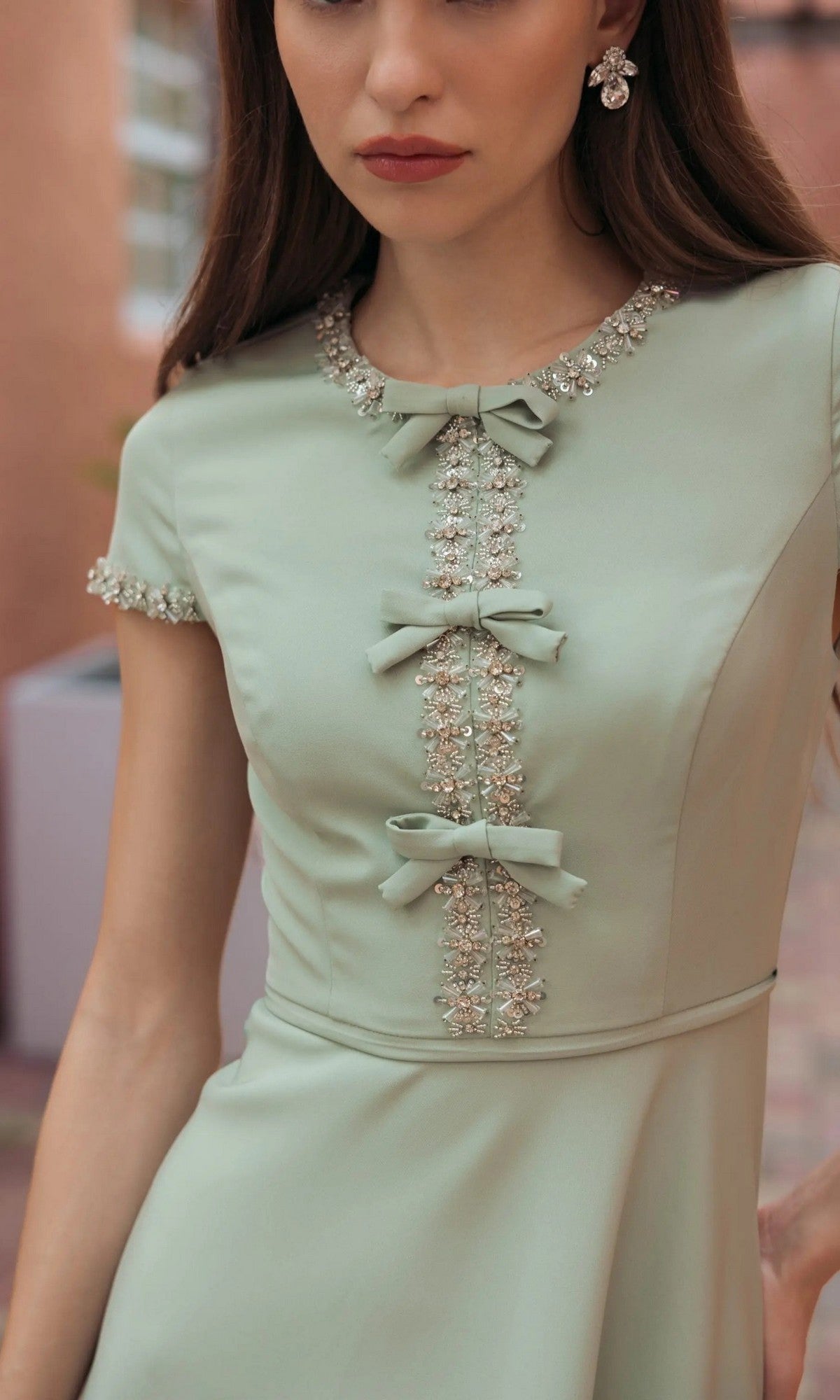 A woman stands outdoors wearing the Midi Party Dress: Mac Duggal 56242—a sage green midi dress featuring short sleeves, three decorative bows, and bead embellishments along the neckline, sleeves, and bodice—making it an elegant choice for a wedding guest ensemble.