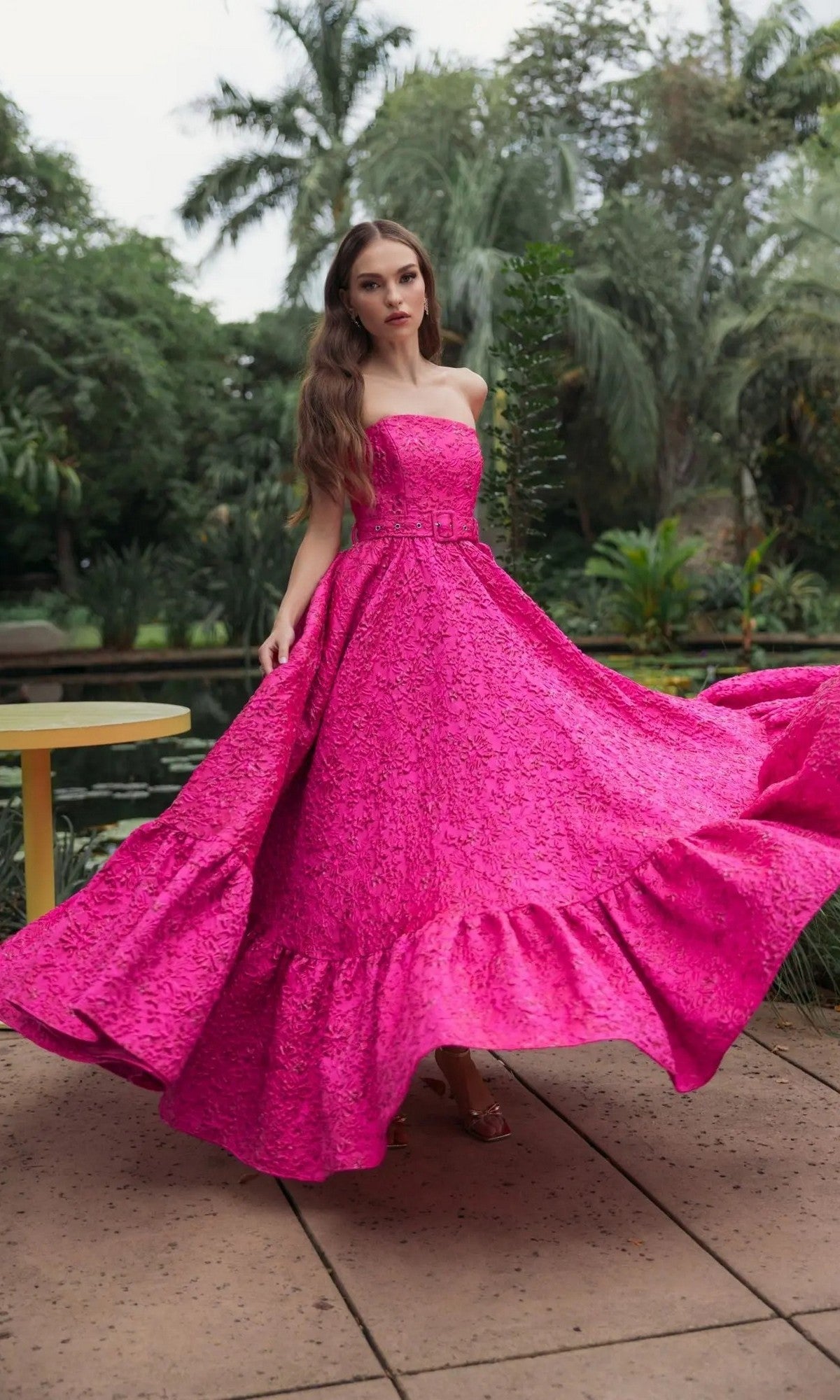 A woman stands outdoors on a patio surrounded by lush greenery, exuding elegance in the Mac Duggal 56277 long formal dress in hot pink, featuring a strapless design with a textured finish and ruffled hem.