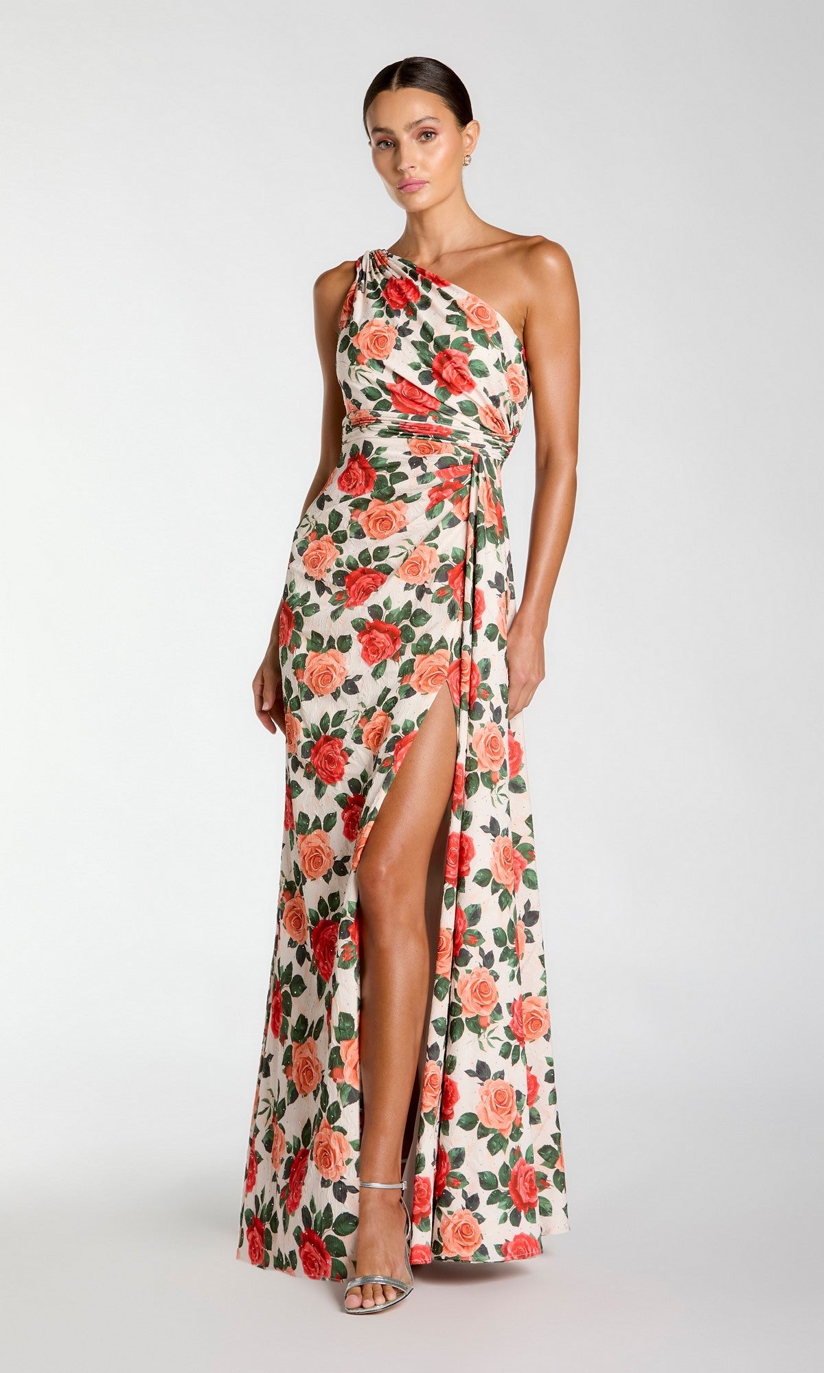 A woman wearing the Long Formal Dress: Mac Duggal 56297 in Red Rose—a floor-length, one-shoulder gown featuring a red and green rose print with a side slit—paired with strappy heels, stands against a plain background.