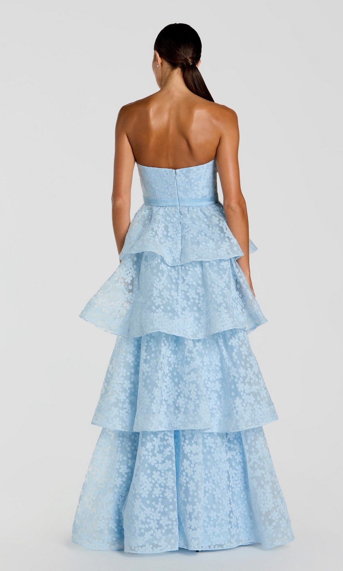 A woman is wearing the Mac Duggal 56298 Long Formal Dress—a strapless, light blue organza gown with a tiered floor-length silhouette and delicate floral pattern—facing away from the camera against a plain background.