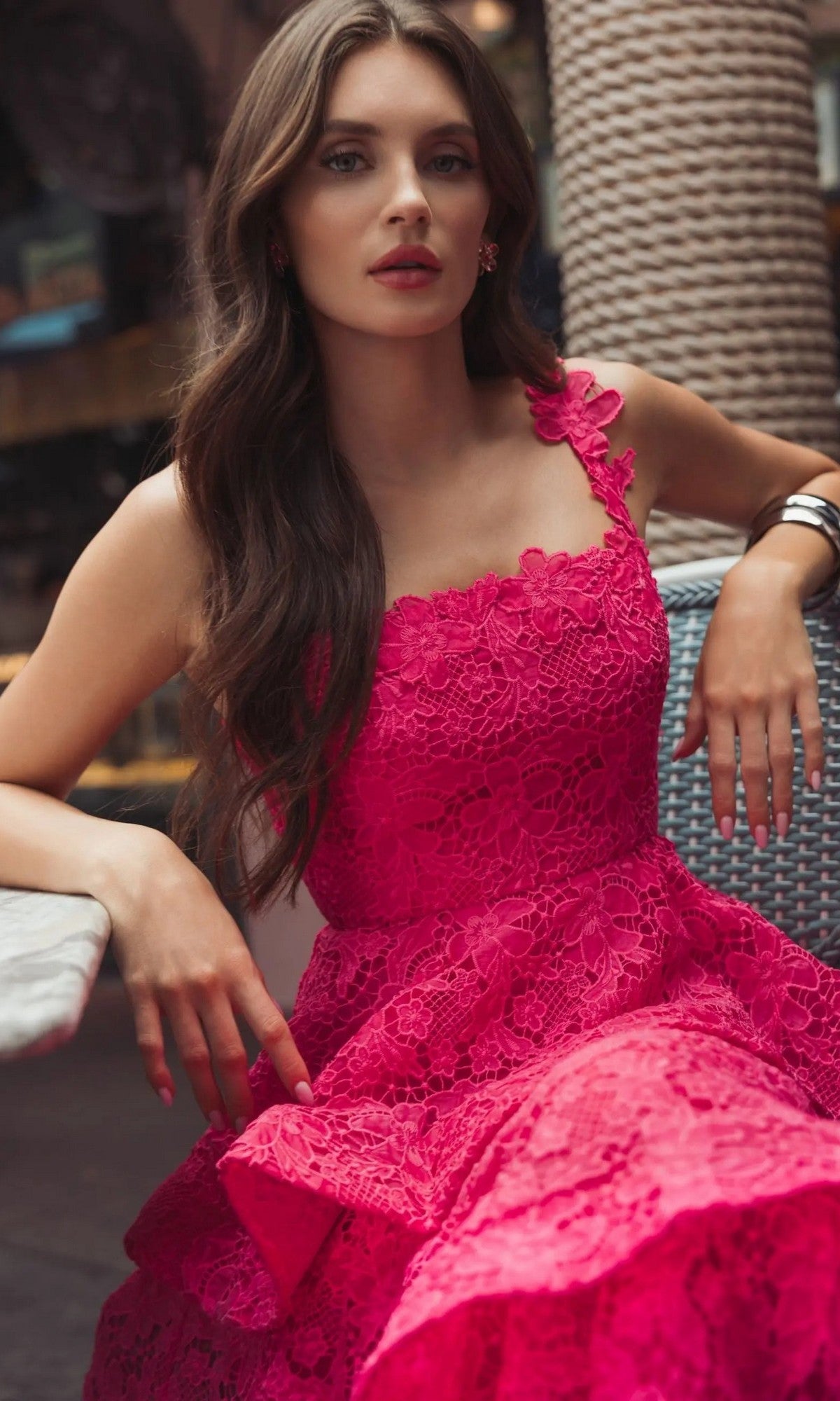A woman with long brown hair sits on a woven chair outdoors, wearing the sleeveless Mac Duggal 56322 Midi Party Dress in hot pink. The lace embroidery of the dress adds an elegant touch to her vibrant look.