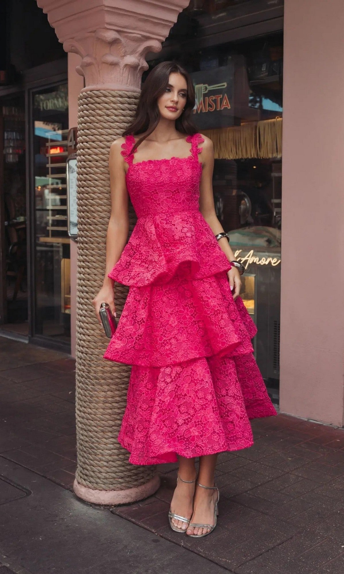 Outdoors, a woman wears the Mac Duggal 56322 Midi Party Dressโa hot pink, sleeveless, tiered lace-embroidered party dressโstyled with strappy heels and a chic clutch.