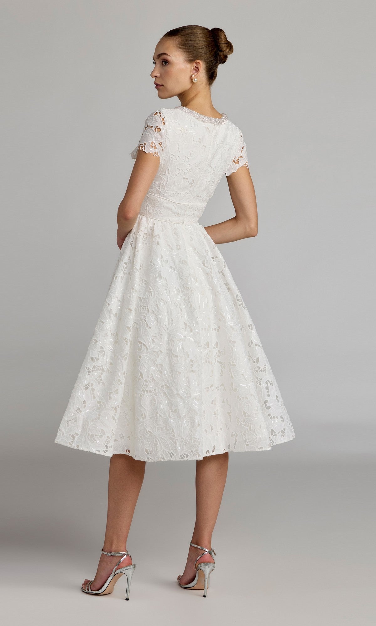 A woman stands with her back turned, looking over her shoulder against a neutral background, wearing the Midi Party Dress: Mac Duggal 56324โa white lace party dress with short sleeves and a tea-length, fit-and-flare midi silhouette. She completes the elegant look with silver high-heeled sandals.