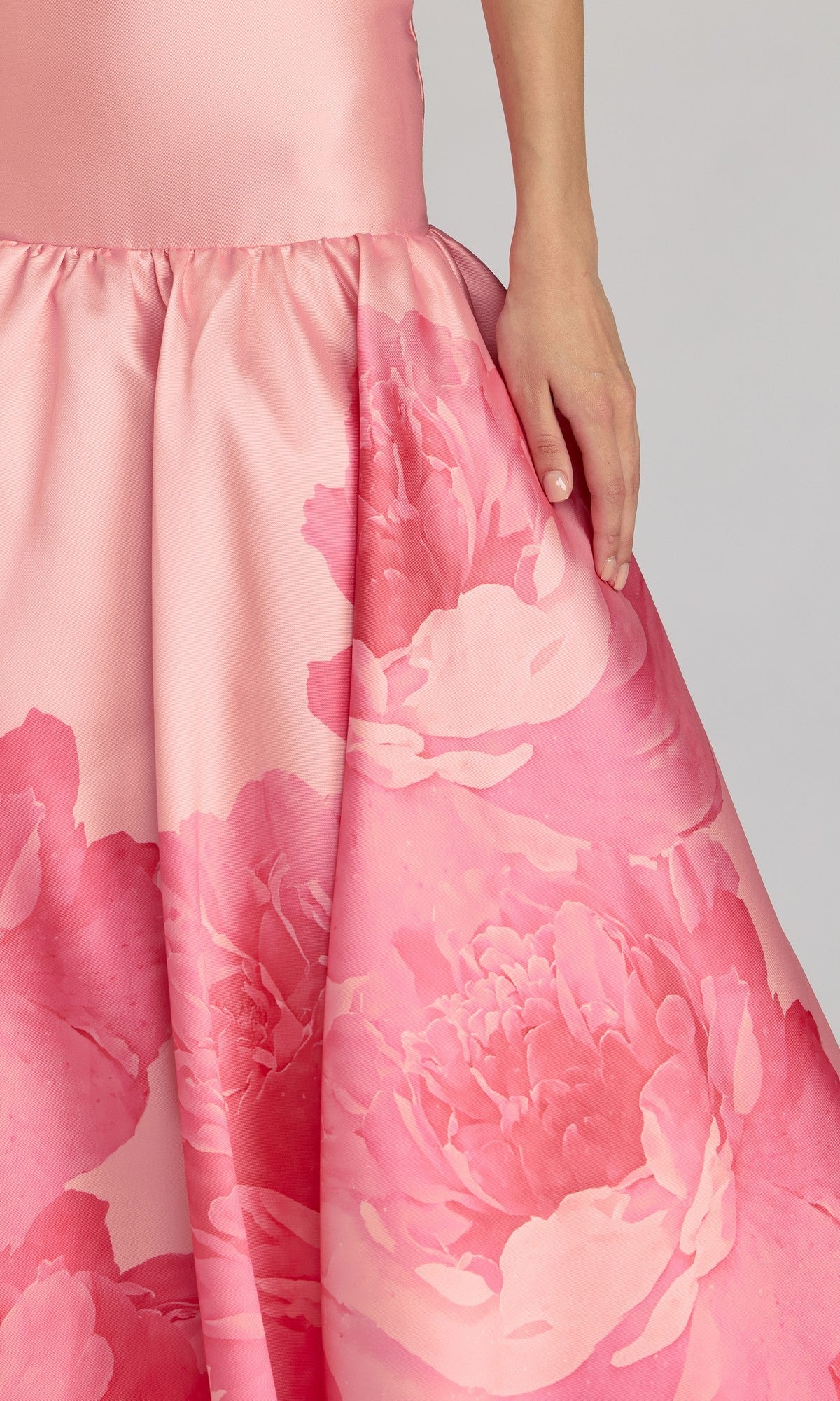 A person is shown from the lower torso down, with one hand gracefully resting on the skirt of the Mac Duggal 56343 Long Formal Dress—a pink satin gown featuring large floral patterns and a Mikado drop waist.