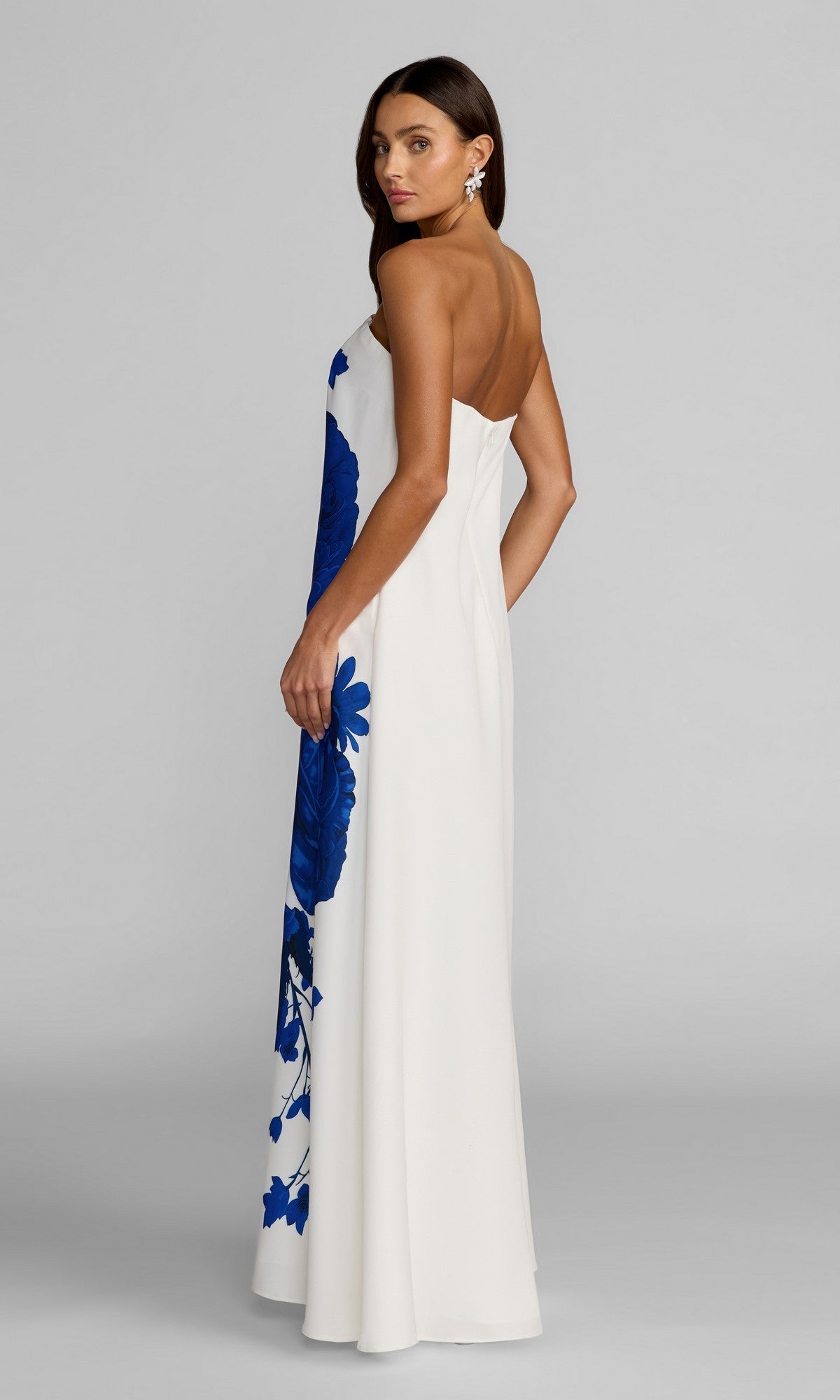 A woman stands against a plain background, looking over her shoulder, wearing the Mac Duggal 56344 long formal dress—a white strapless gown adorned with a bold blue floral print.