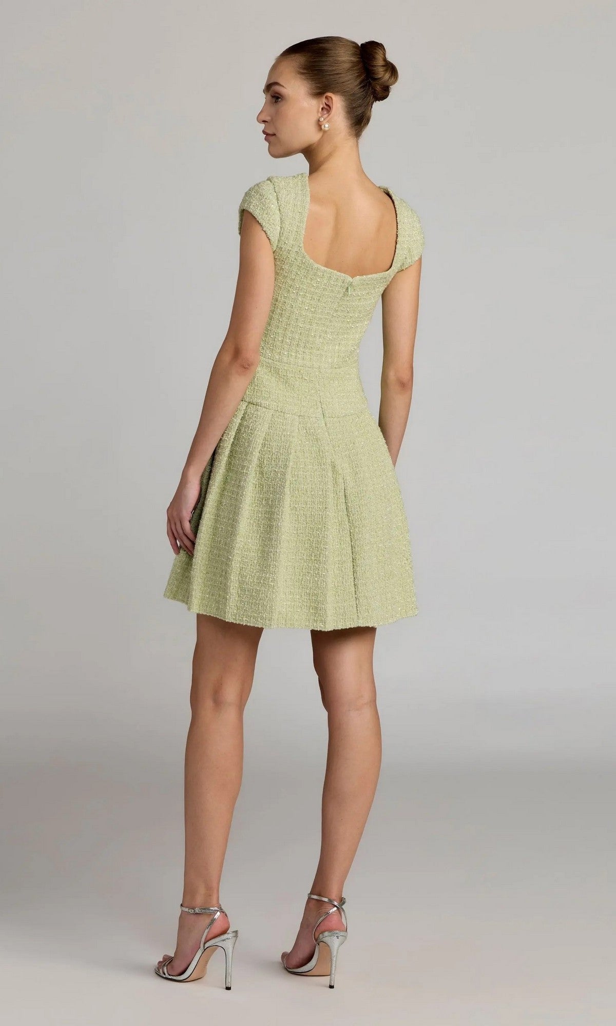 A woman stands with her back to the camera, wearing the Mac Duggal 56373 Short Casual Dress in light green boucle with cap sleeves and textured fabric, paired with silver high-heeled sandals. Her hair is styled in a bun against a plain light background.