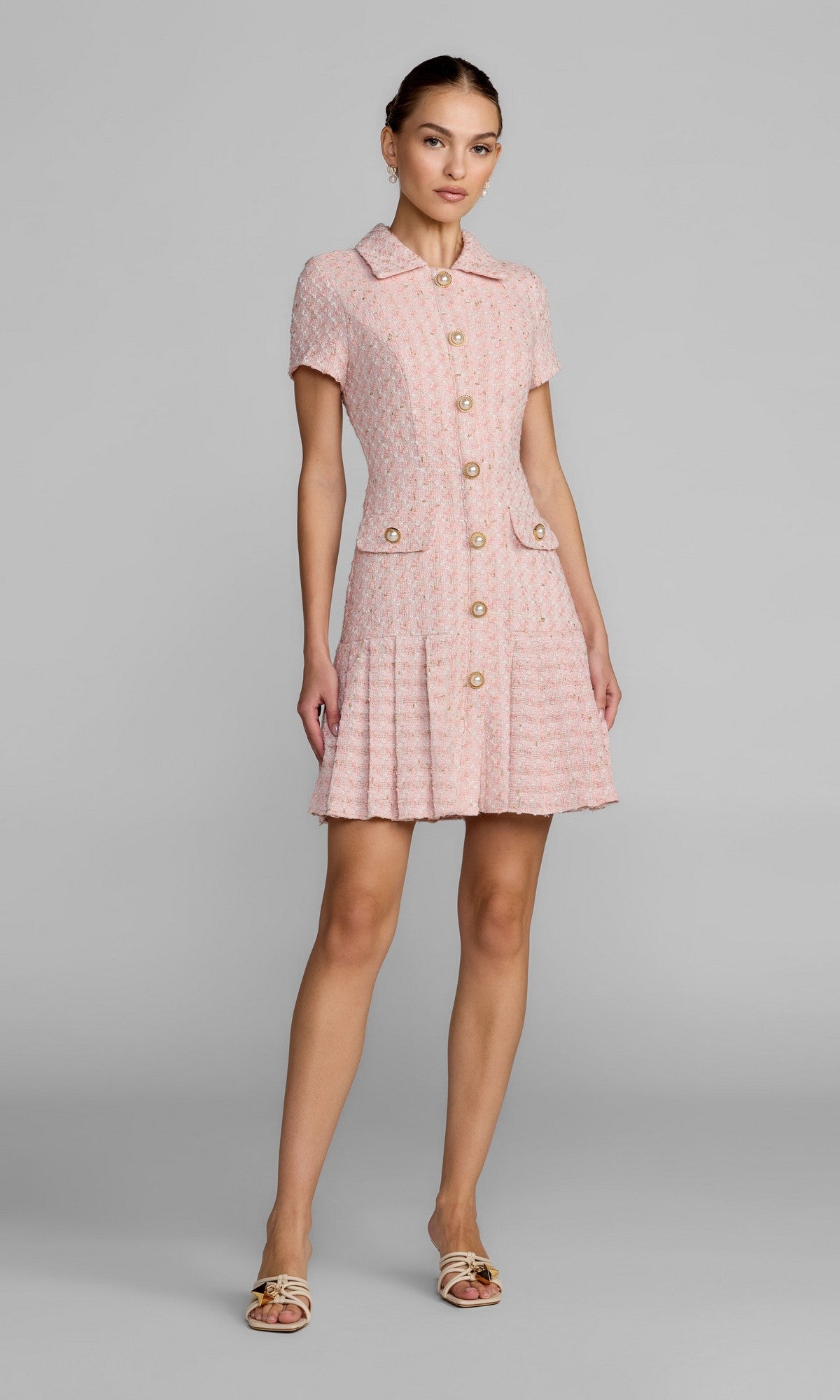 A woman stands in a studio wearing the Mac Duggal 56379 Short Party Dress in pink, featuring short sleeves, a pleated A-line silhouette, front pockets, and a textured button-down design, paired with open-toe heels.