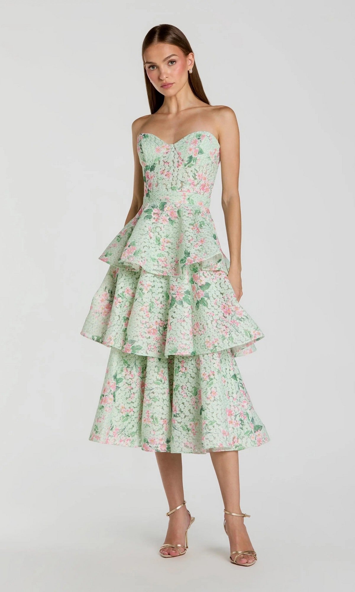A woman stands against a plain background wearing the Midi Wedding Guest Dress: Mac Duggal 56389—a strapless, pastel floral-printed net dress featuring a fitted bodice and tiered ruffle skirt—paired with strappy heels.