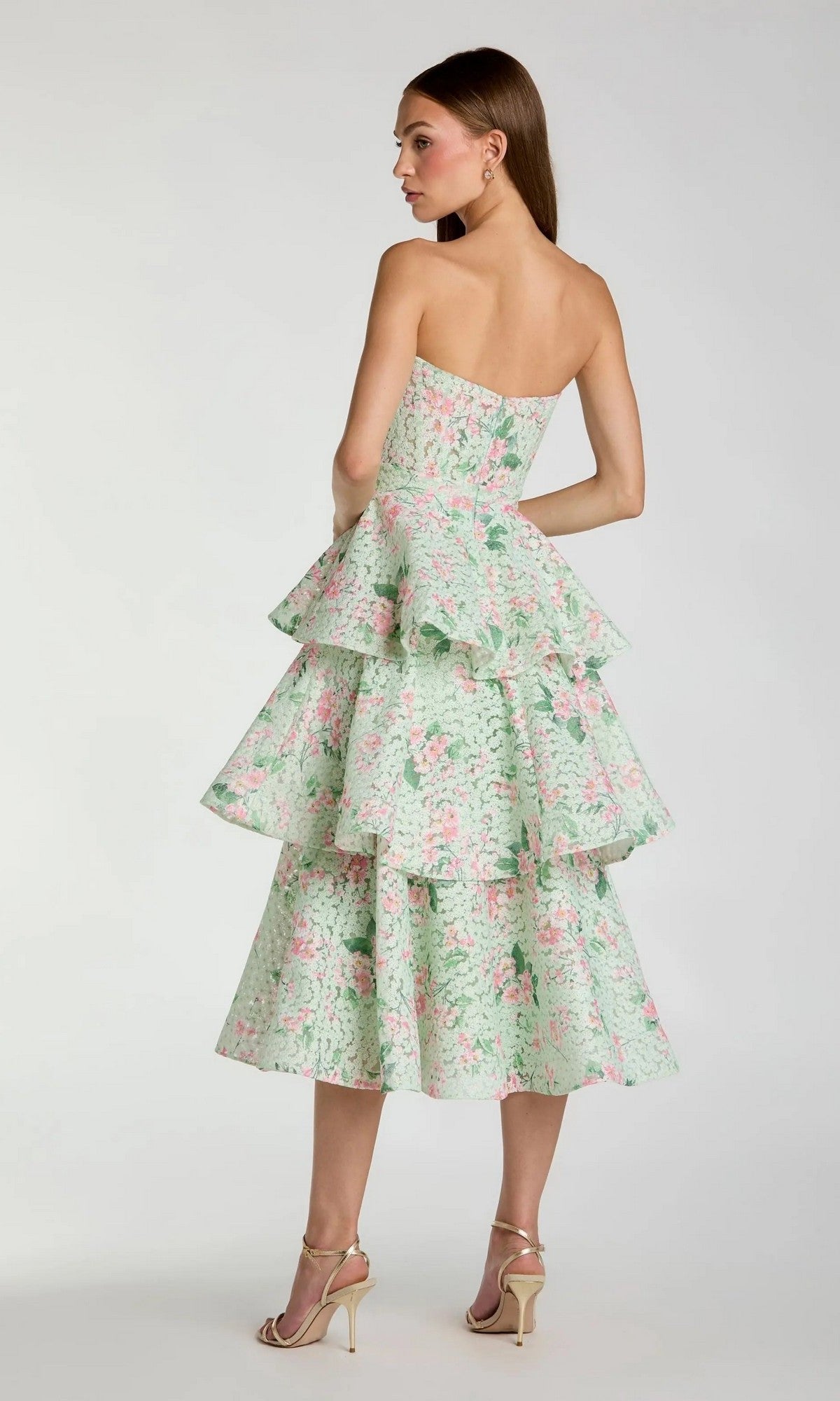 A woman wearing the Mac Duggal 56389 Midi Wedding Guest Dress—a strapless, tiered net dress with a pastel floral print—and strappy heels stands facing away against a plain background.