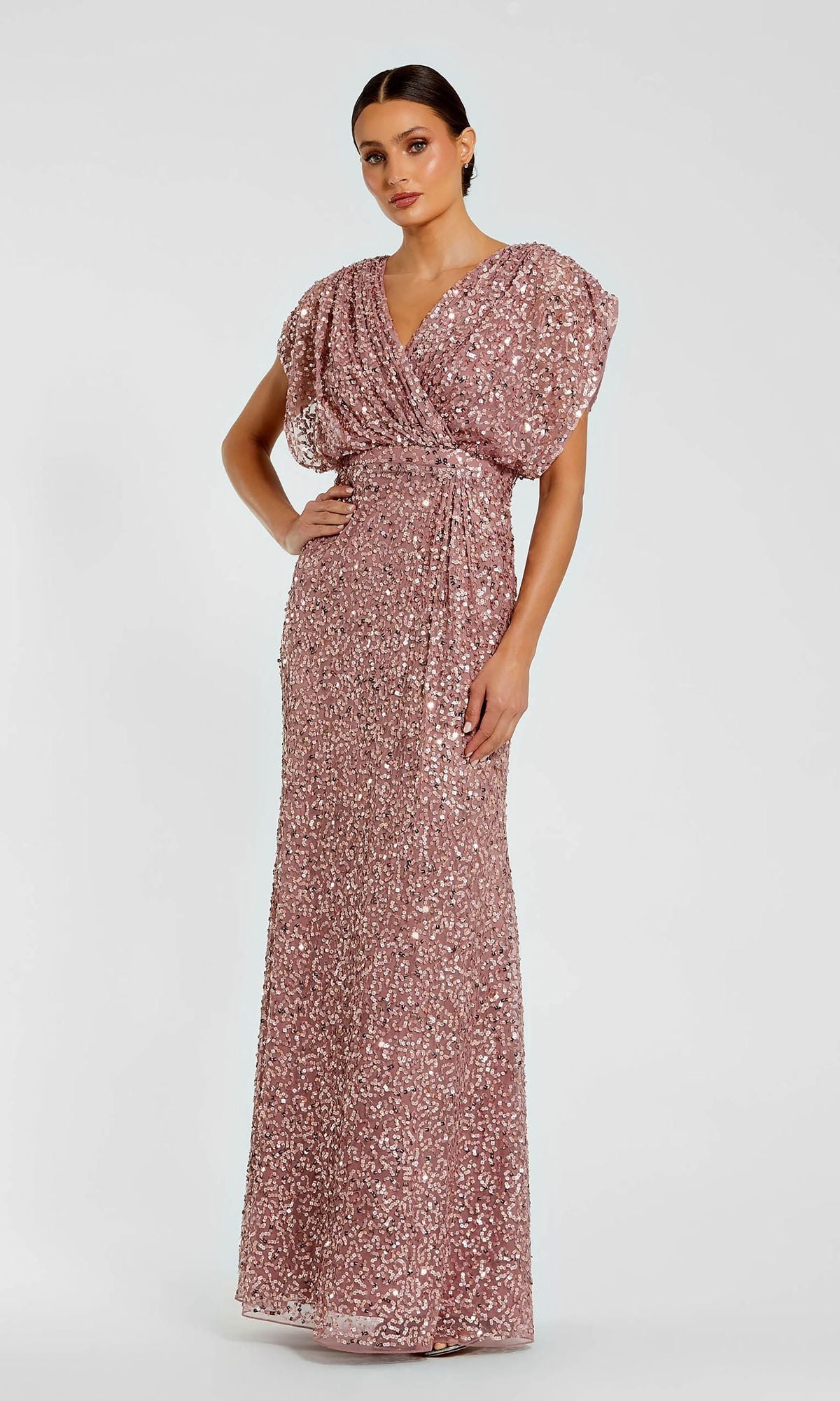 A woman stands against a white background wearing the Mac Duggal 5640 Short Sleeve Long Sequin Formal Dress in rose gold, featuring a draped V-neckline and fitted silhouette.