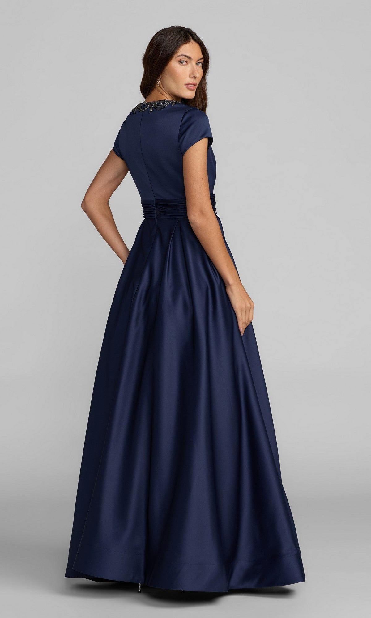 A woman in the Mac Duggal 56401 Long Formal Dress—a dark blue, floor-length satin gown with short sleeves and a fitted waist—stands against a plain background, looking over her shoulder; the elegant silhouette embodies classic Mac Duggal sophistication.