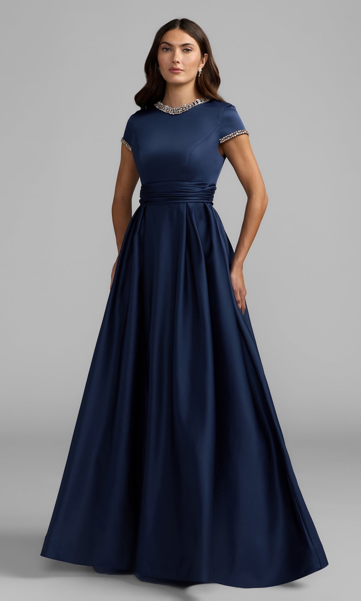 A woman stands wearing the Long Formal Dress: Mac Duggal 56402 in Midnight, a floor-length gown featuring short sleeves, a fitted bodice, jeweled collar, and waistband, as she poses against a plain gray background.