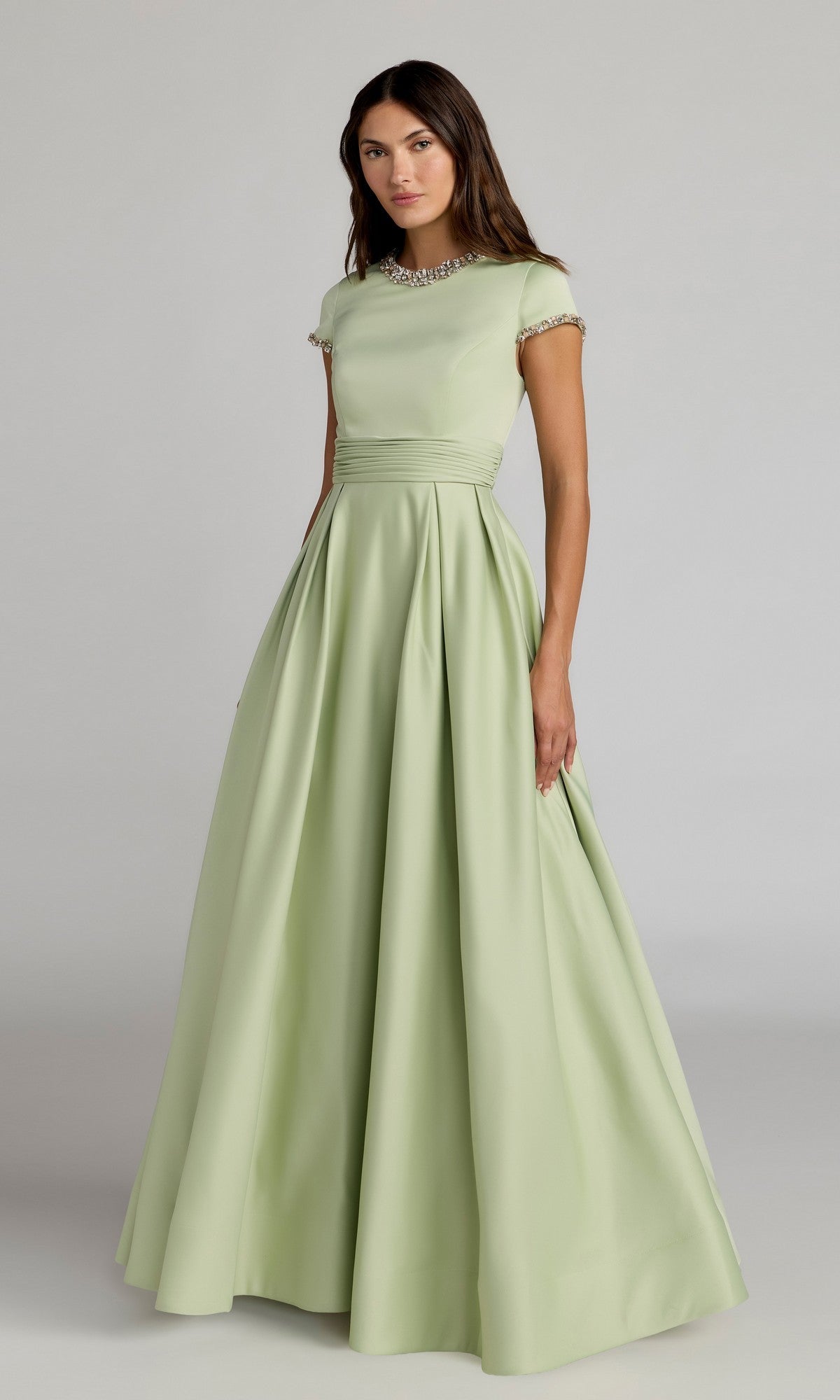 A woman stands wearing the Long Formal Dress: Mac Duggal 56402 in sage—a floor-length formal gown with short sleeves, a fitted bodice, jeweled neckline, and a full luminous satin skirt. The elegant silhouette captures the timeless sophistication of Mac Duggal against a plain background.