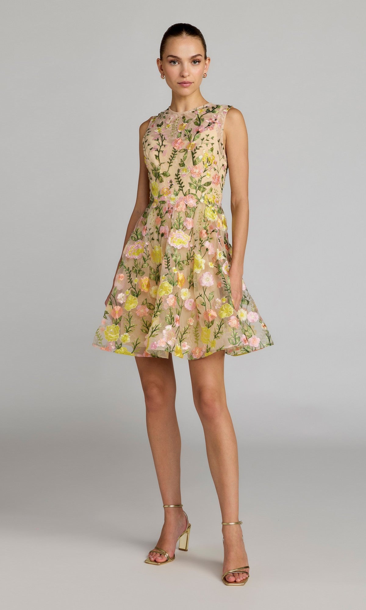 A woman wears the Mac Duggal 56409 Short Party Dress in Yellow Multi, featuring a sleeveless design, knee-length hem, floral pattern, and an A-line fit-and-flare silhouette. She pairs it with gold heels and stands against a plain light gray background.