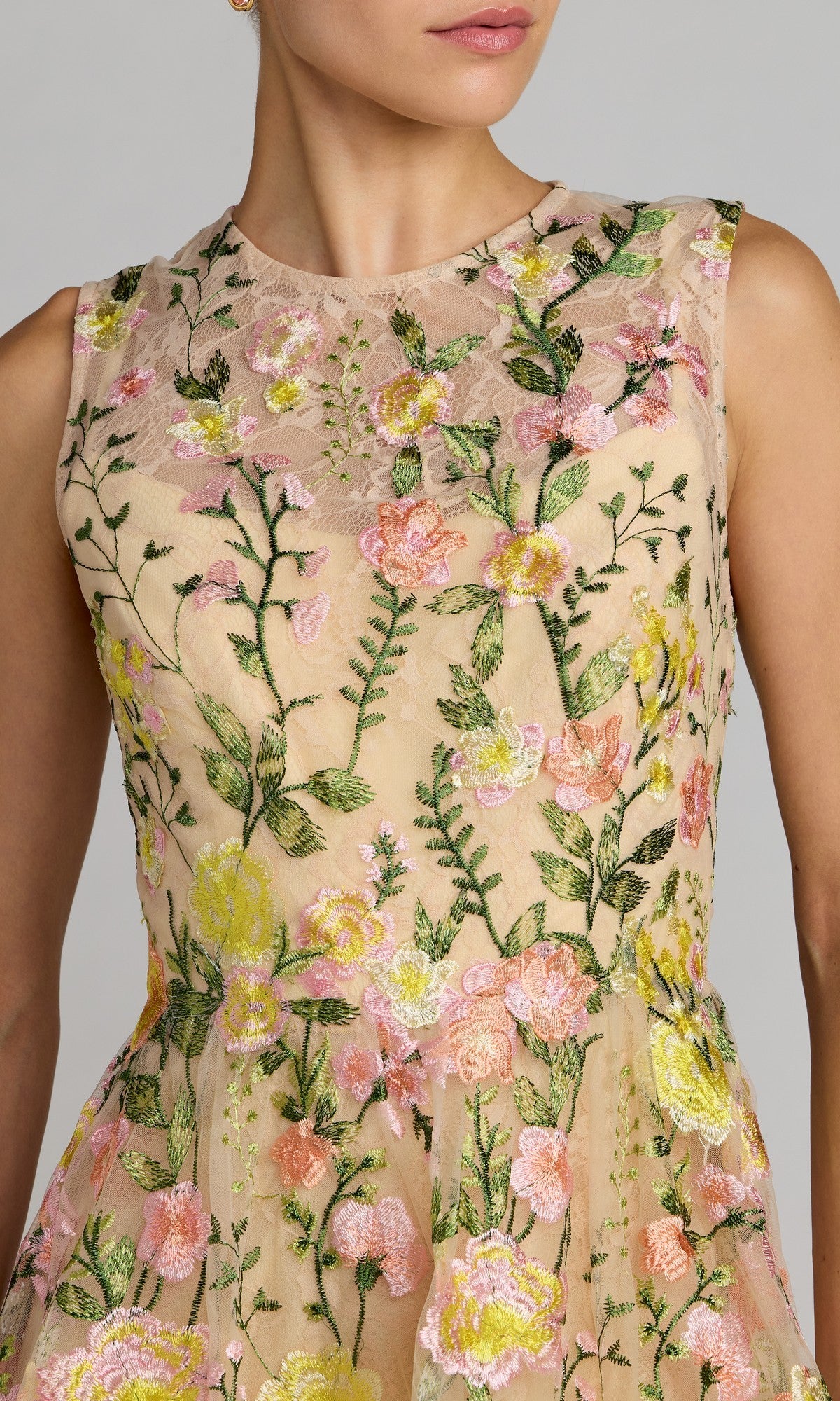 A woman wearing the Mac Duggal 56409 short party dress, a sleeveless mini style adorned with intricate pink, yellow, and green floral embroidery on sheer lace. This dress features a sheer high neckline and a flattering fit-and-flare silhouette that enhances the elegance of its vibrant embroidered details.